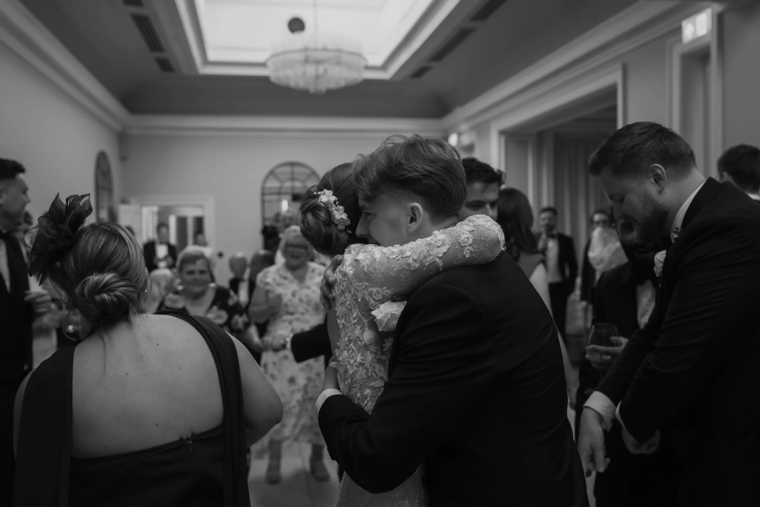 A black and white photo of a wedding reception with people dancing and socializing indoors. A couple is hugging in the center, the woman in a lace dress and floral hair accessory, the man in a black suit.