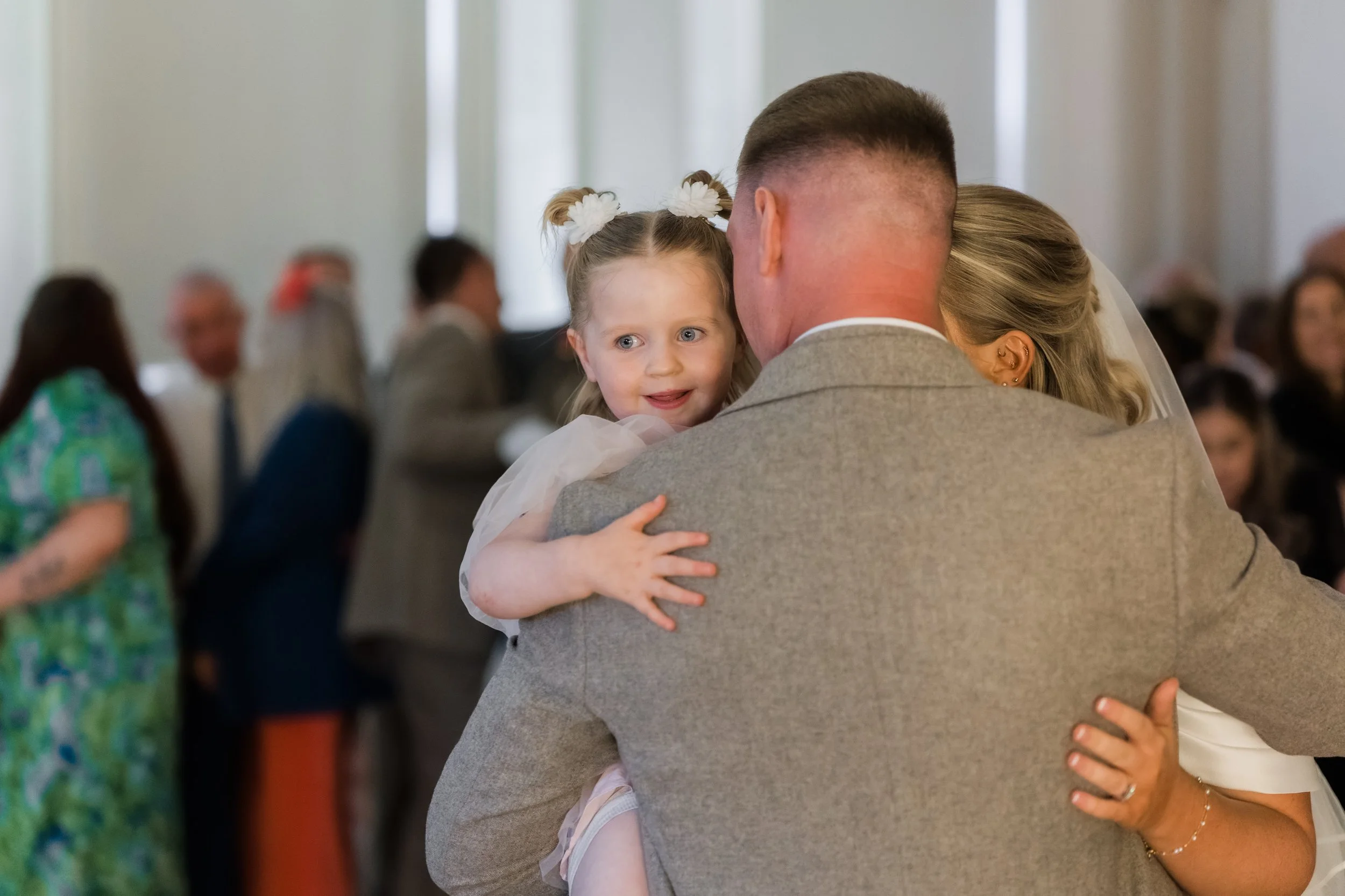 A man and woman embrace a young girl at a wedding reception, with other guests in the background.