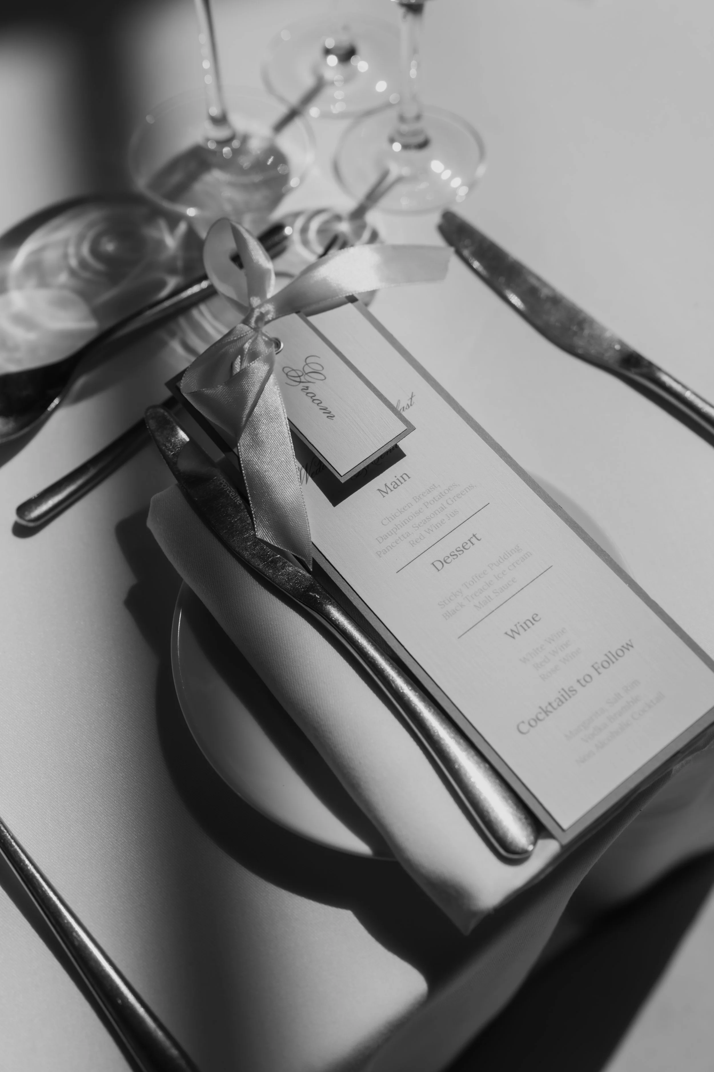 A wedding menu placed on a tray with a ribbon, three empty wine glasses in the background, a knife, and a fork.