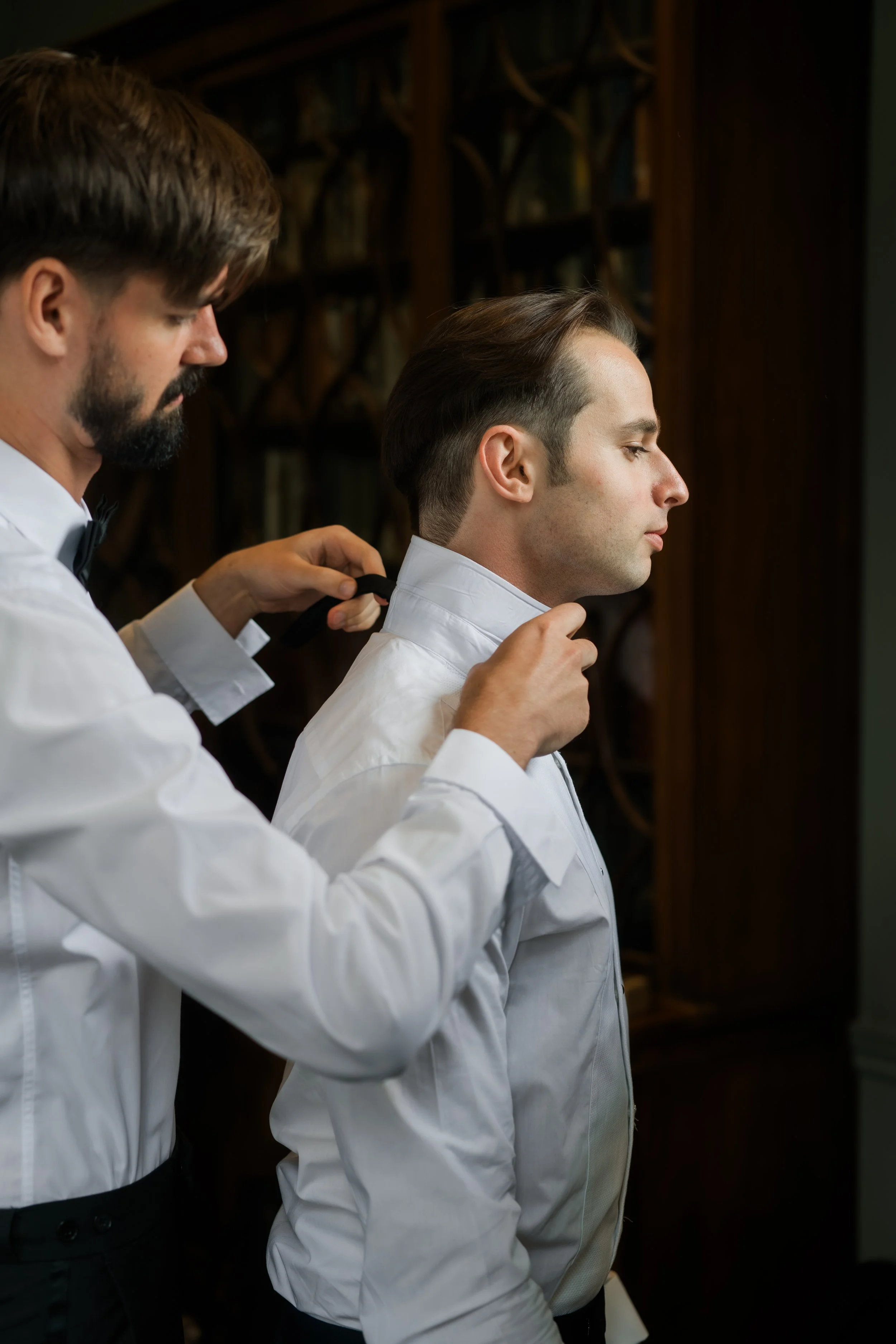 A man in a white dress shirt standing with a serious expression as another man assists with fastening his collar before a formal event.