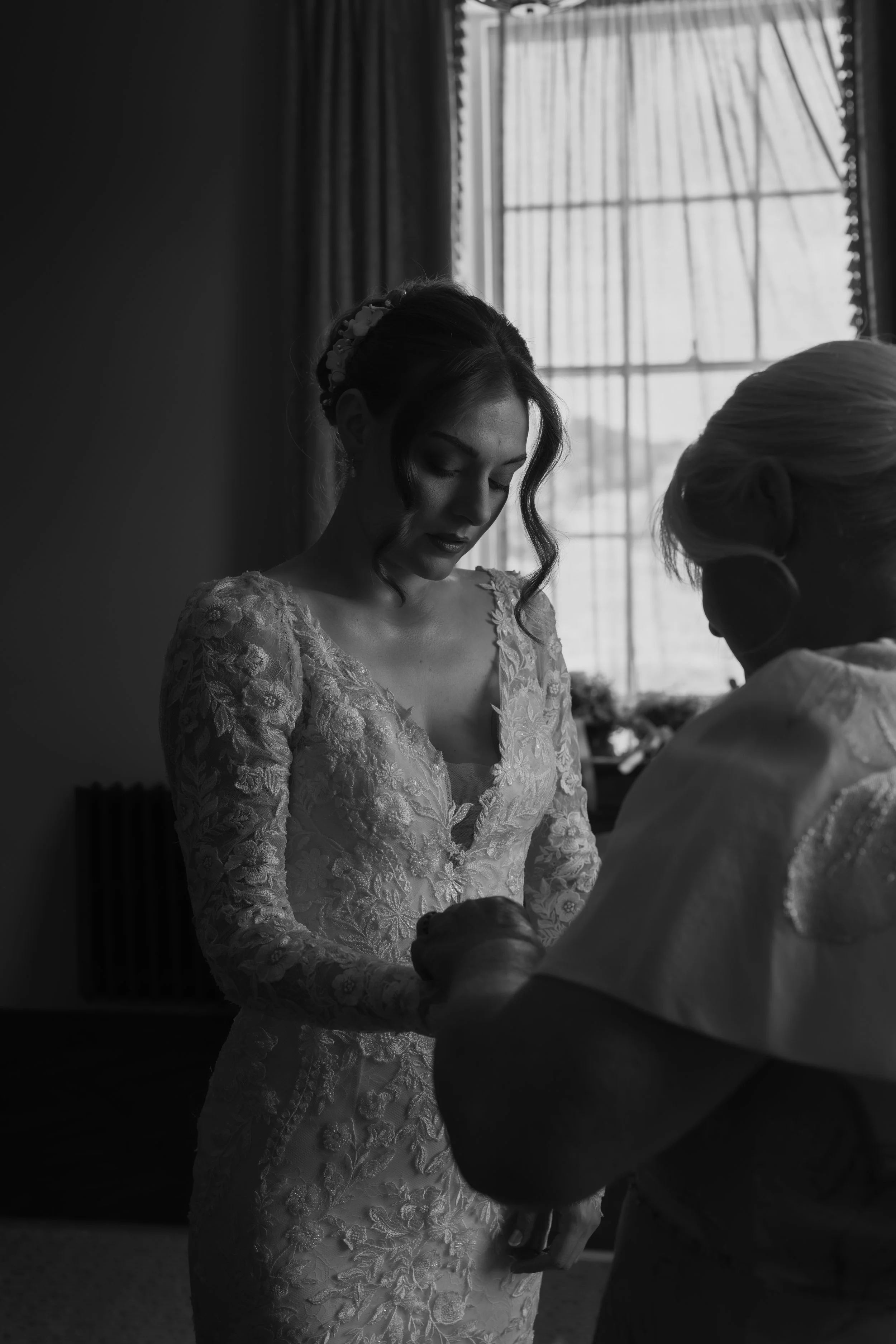 A bride in a lace wedding dress with floral details and a flower headband standing by a window, holding hands with an older woman, possibly during a wedding moment.