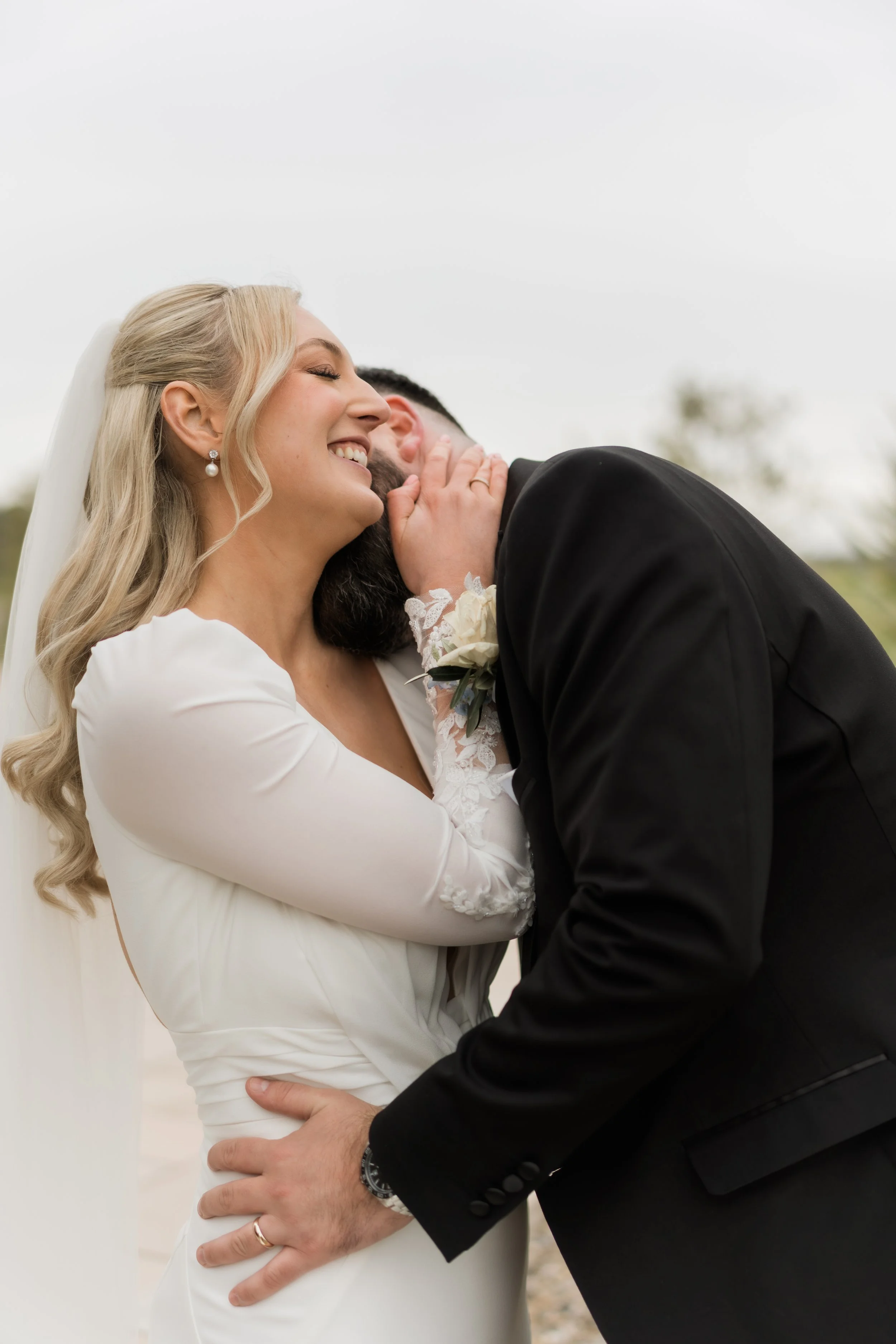 A bride and groom share an intimate moment, with the groom whispering in the bride's ear as she smiles. The bride wears a white wedding dress and pearl earrings, while the groom wears a black suit. The background is outdoors with blurred trees and sk