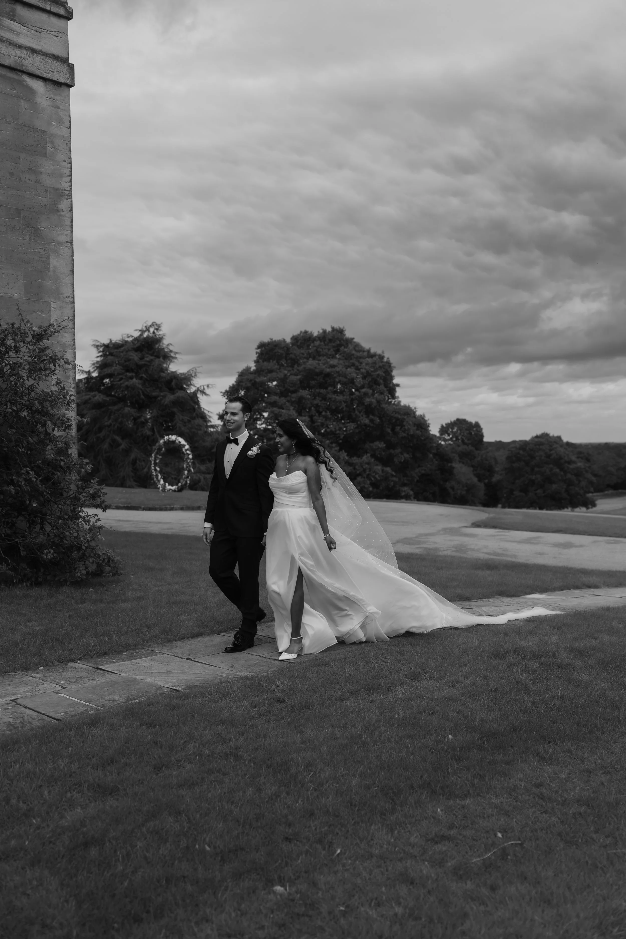 A black and white photograph of a bride and groom walking outside on a cloudy day, the bride wearing a strapless wedding gown and veil, and the groom in a tuxedo with a bowtie, near a large stone building and a grassy area with trees in the backgroun