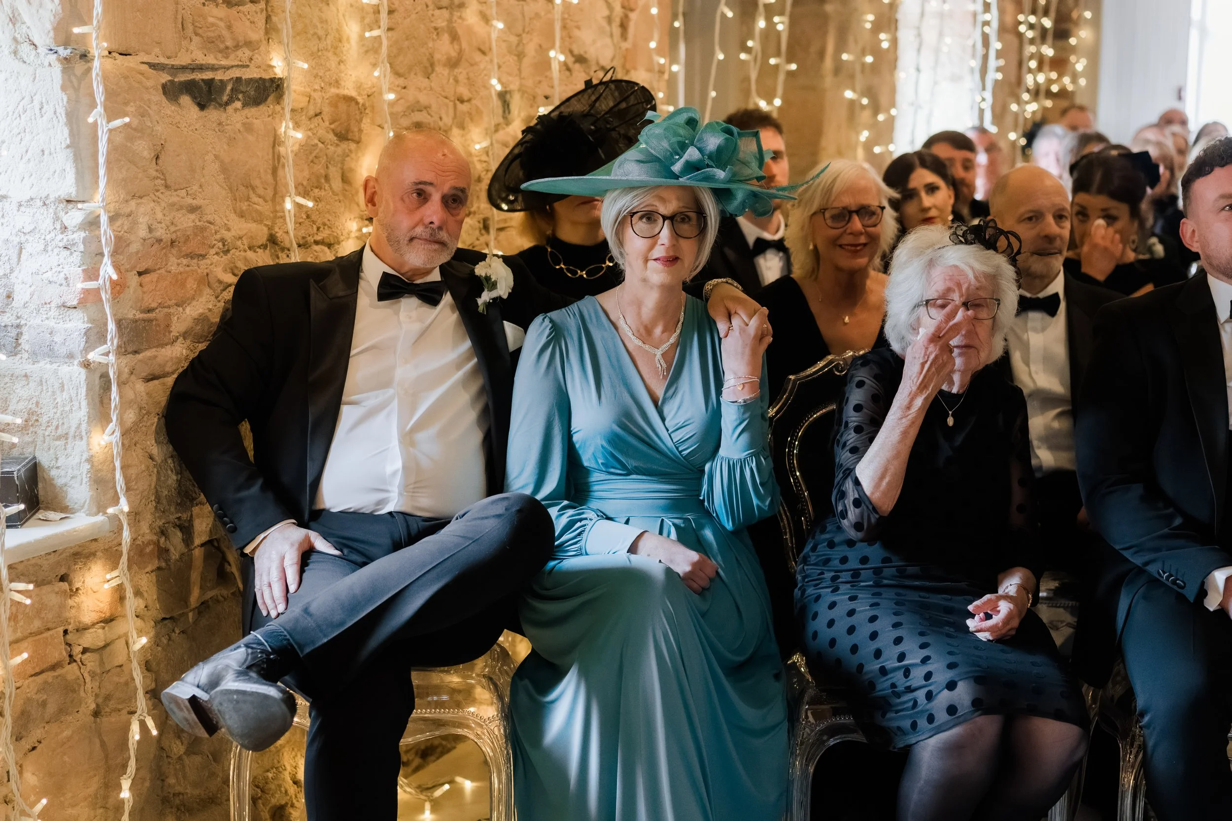 Guests sitting at a formal event, with string lights and a stone wall in the background. A woman in a bright blue dress and large teal hat with black netting is seated in the center, flanked by older women and men in tuxedos.