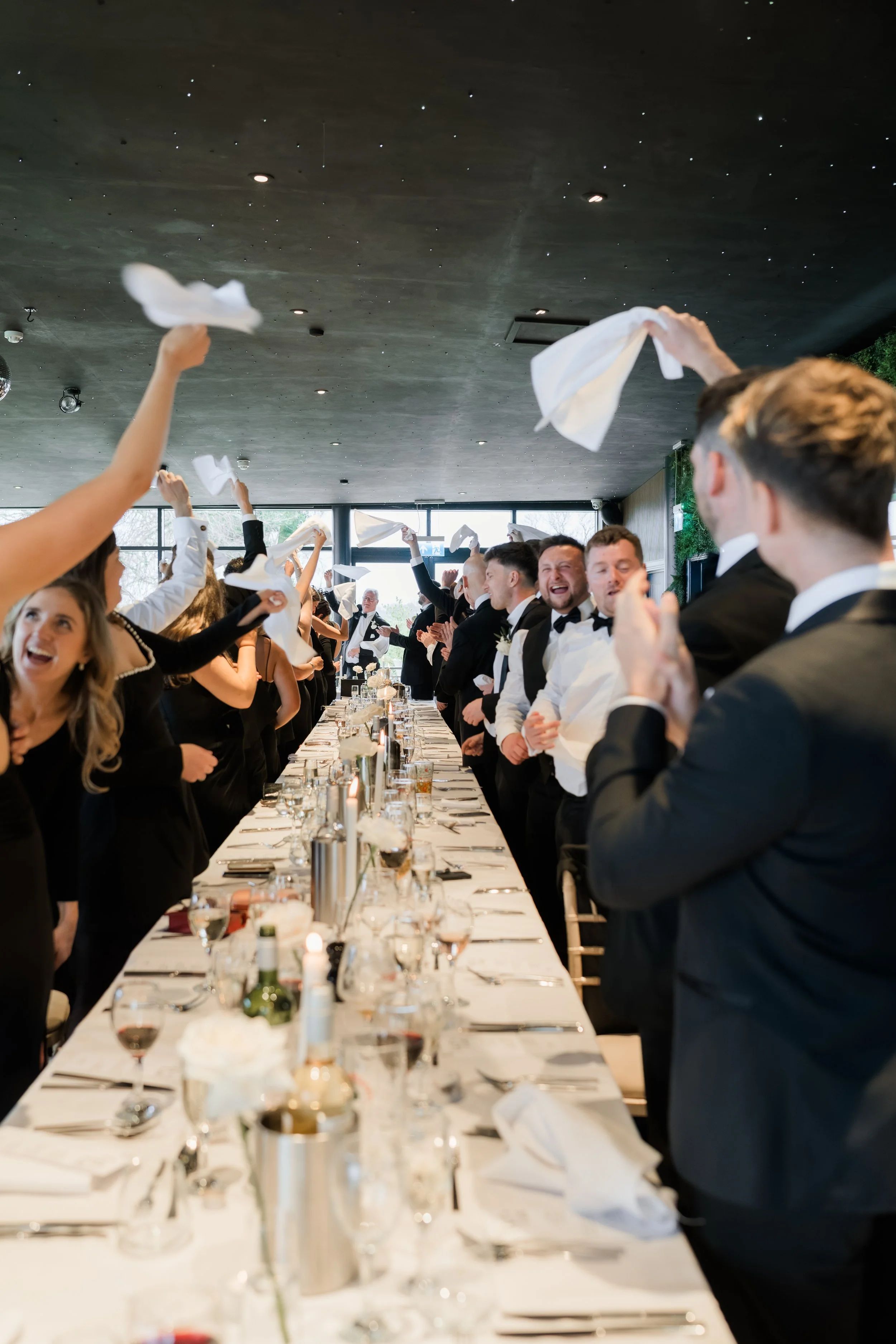People at a wedding reception celebrating by tossing white napkins in the air at a long decorated banquet table.