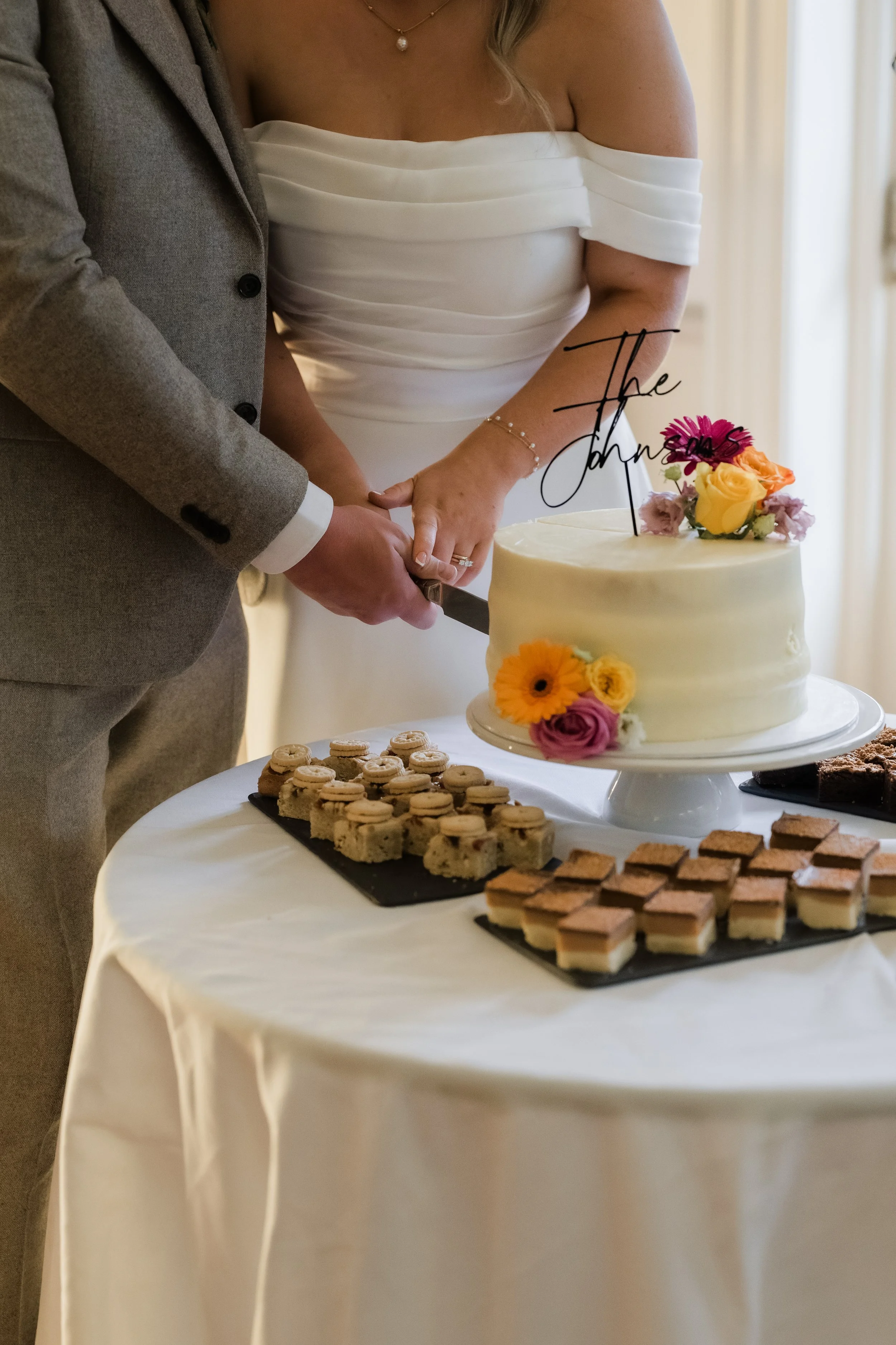 A bride and groom are cutting a wedding cake together at their wedding reception. The cake is decorated with flowers and a topper that reads 'The'.