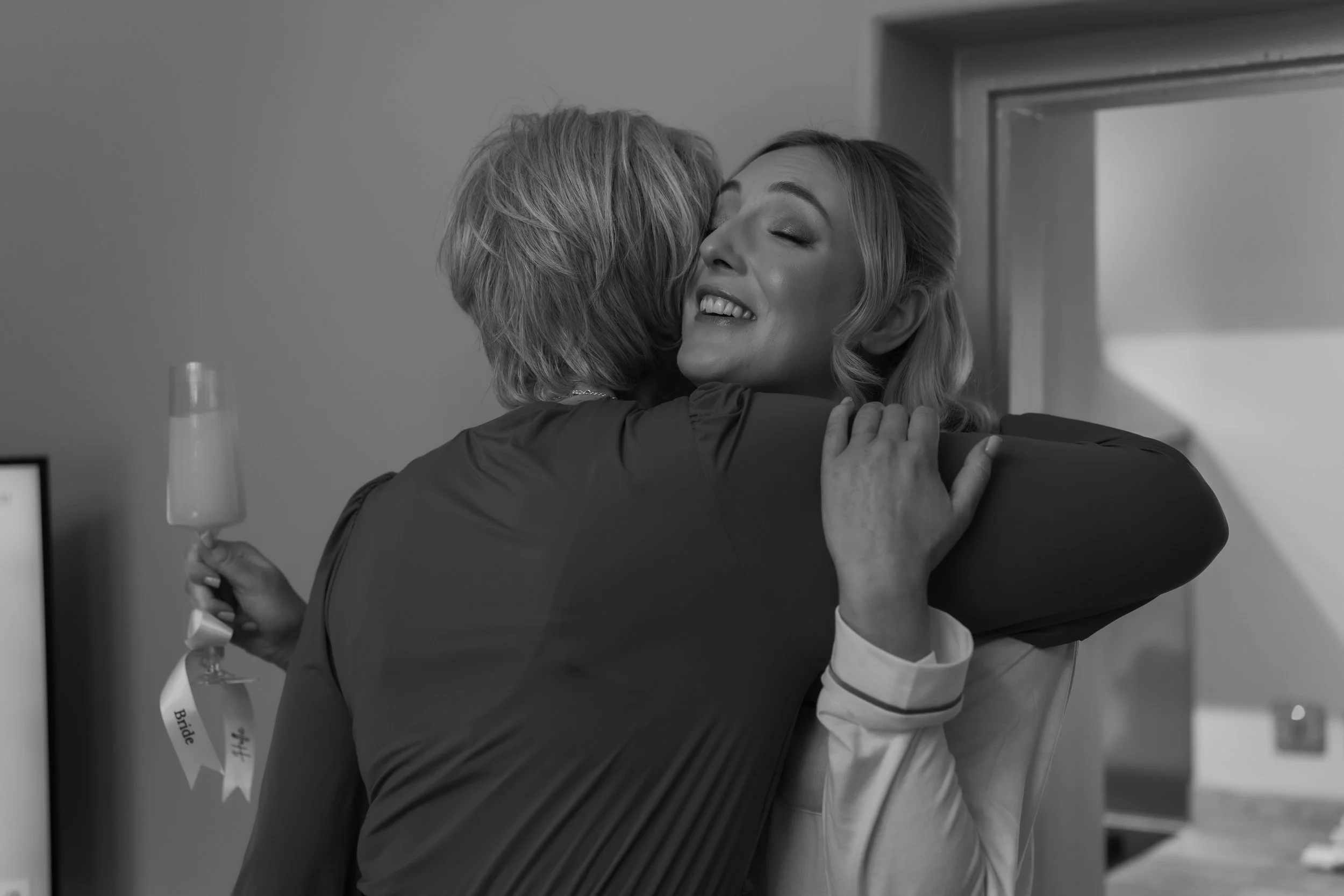 A woman smiling and embracing another person in a hug, holding a champagne flute, in a joyful moment.