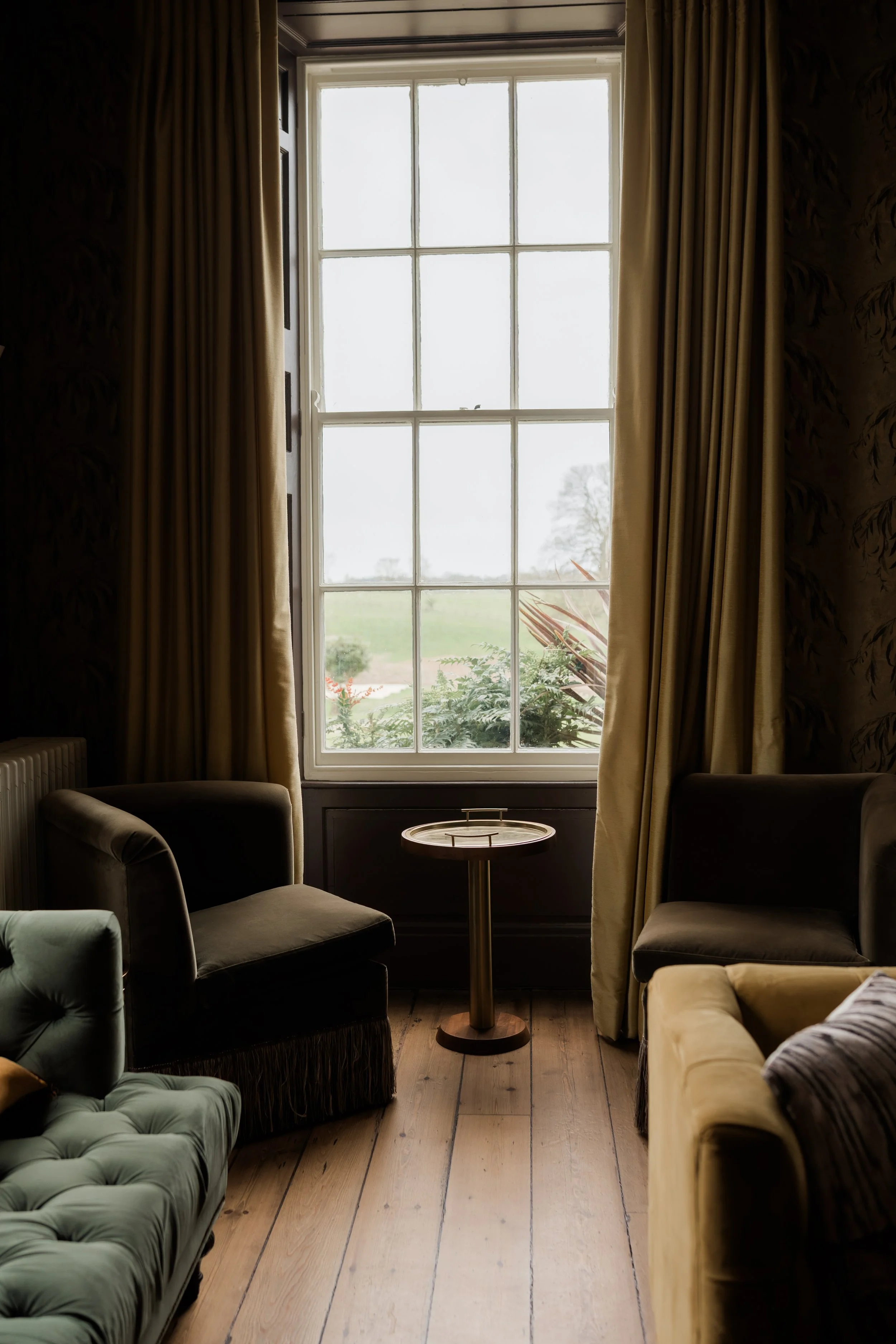 Interior view of a cozy living room with a large window, beige curtains, and vintage-style chairs around a small round table, overlooking a garden.