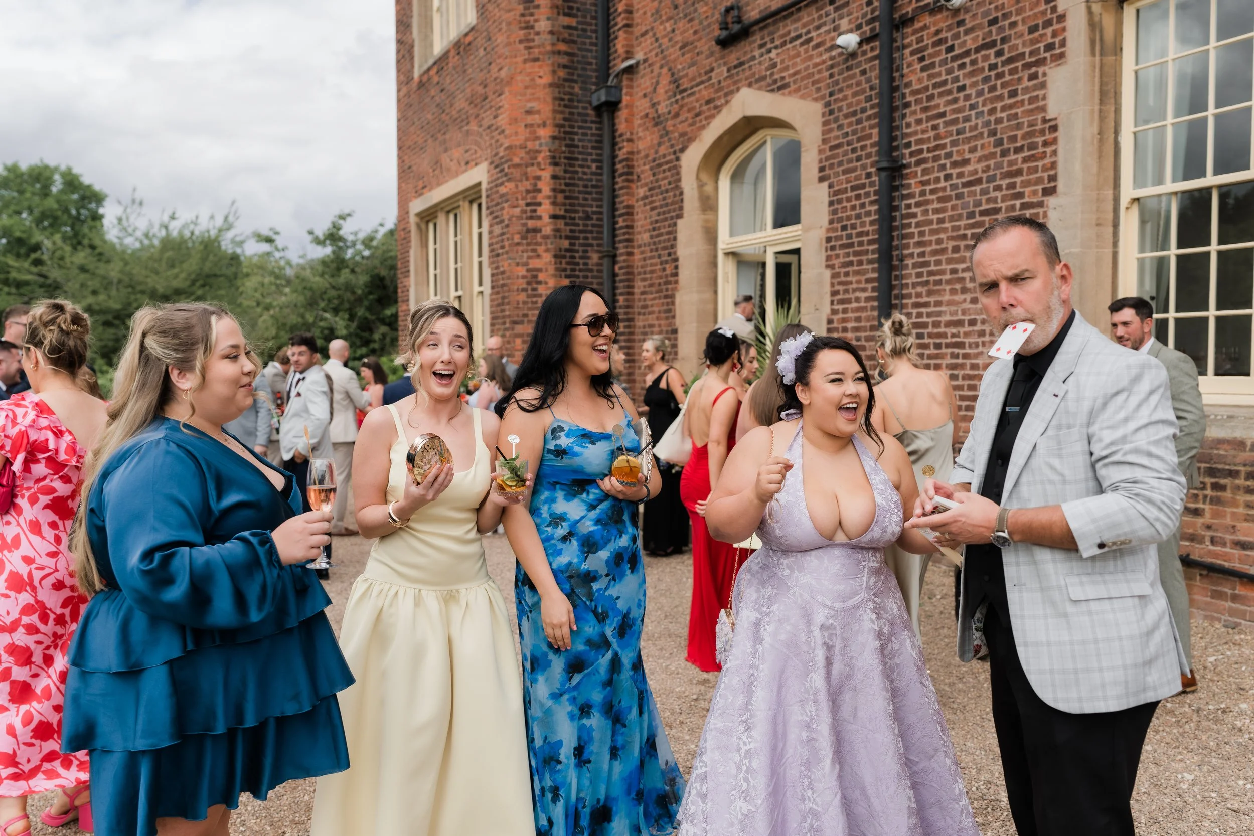 Group of women and a man at a wedding reception outdoors, chatting and laughing. The women are holding drinks and wearing colorful dresses; the man has a playing card in his mouth and is dressed in a suit.