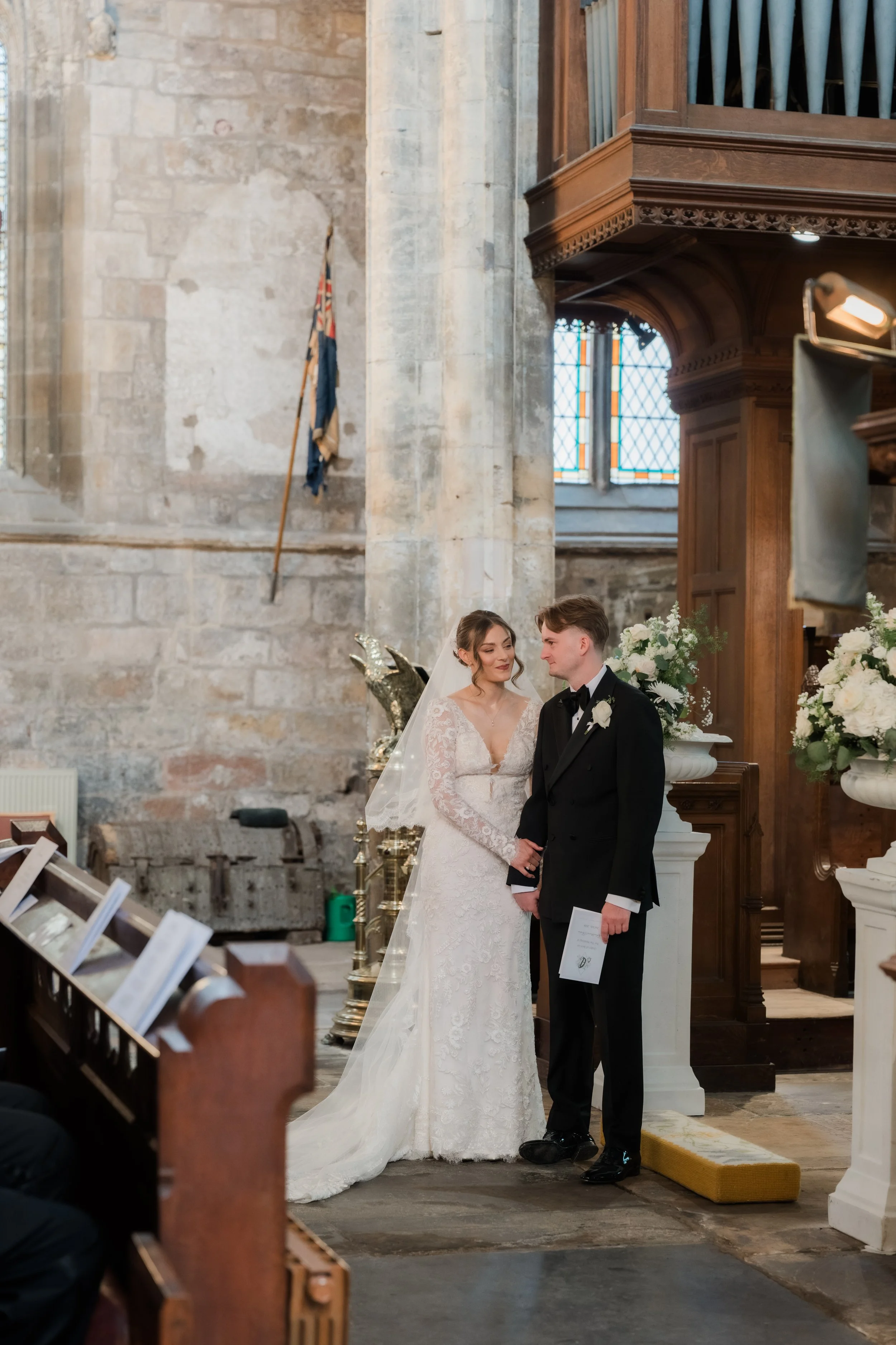 A bride and groom standing together during a wedding ceremony inside a church with stone walls, stained glass windows, and wooden pews.