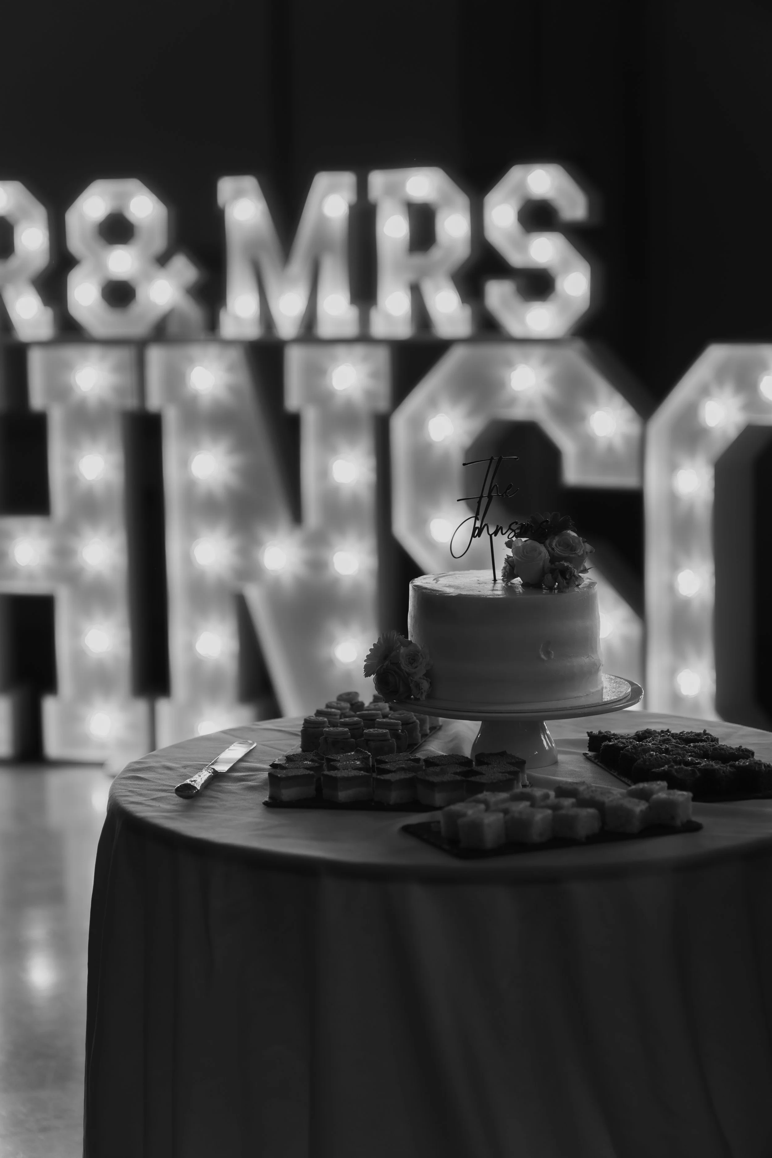 A wedding cake on a table with desserts, with illuminated marquee signs in the background reading '& MRS' and 'THE'.