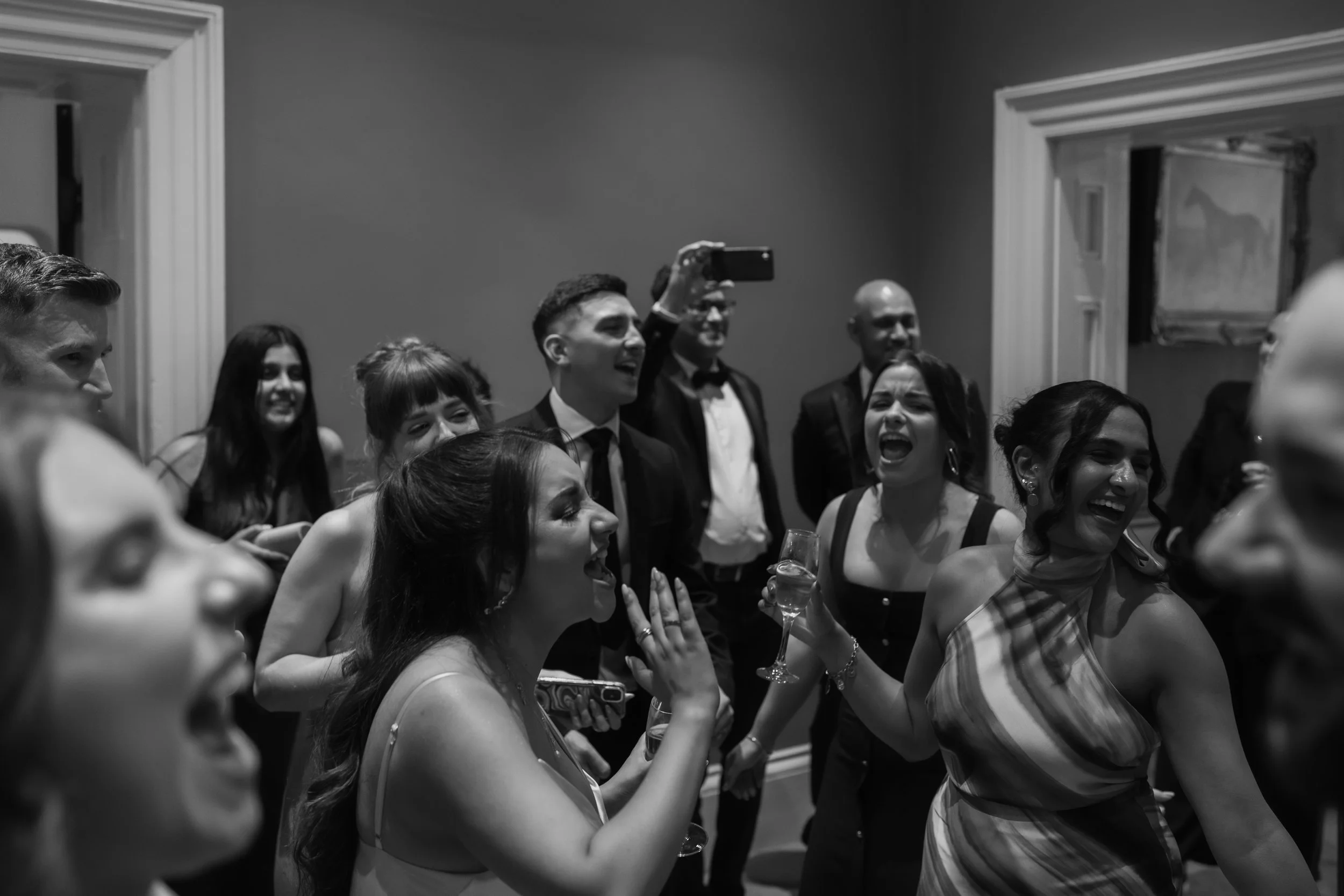 People laughing and smiling at a party, some holding glasses of wine, in an indoor setting.
