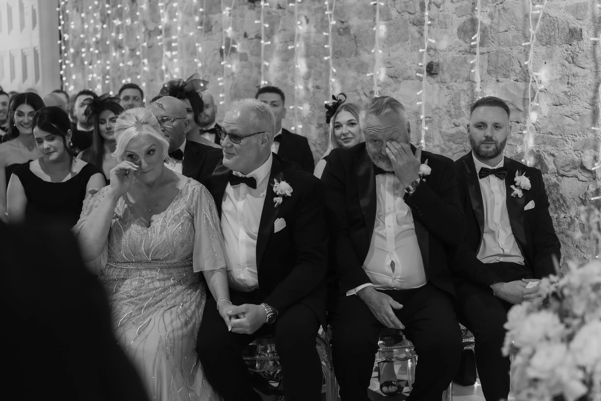 Black-and-white photograph of a wedding ceremony with people seated in rows. The front row includes a woman wiping a tear, an older man holding hands with her, another older man wiping his eye, and a young man with a beard. The background has string 