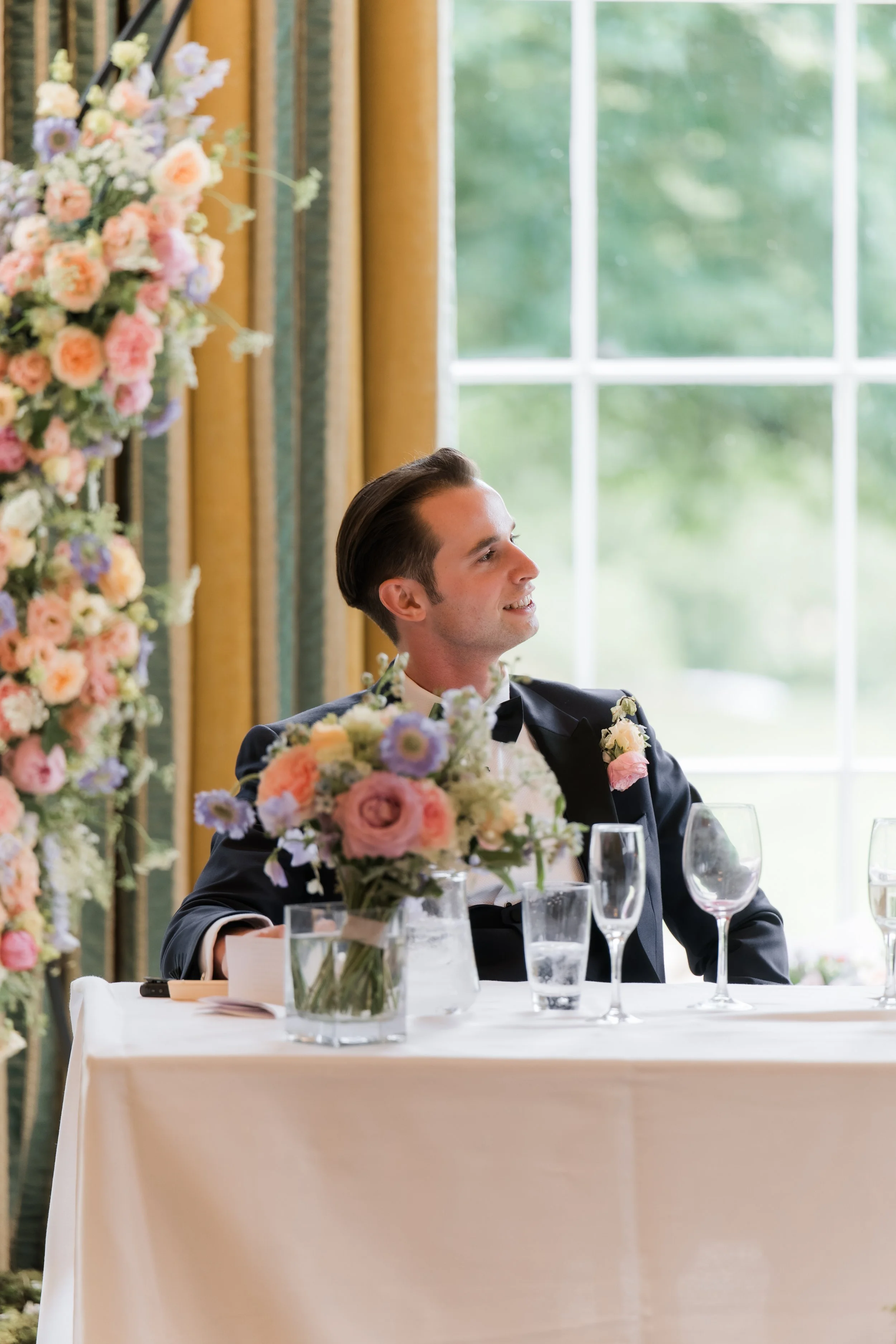 Young man in tuxedo sitting at a table during a wedding reception, decorated with floral arrangements and surrounded by empty wine glasses, with a large window and lush greenery outside.