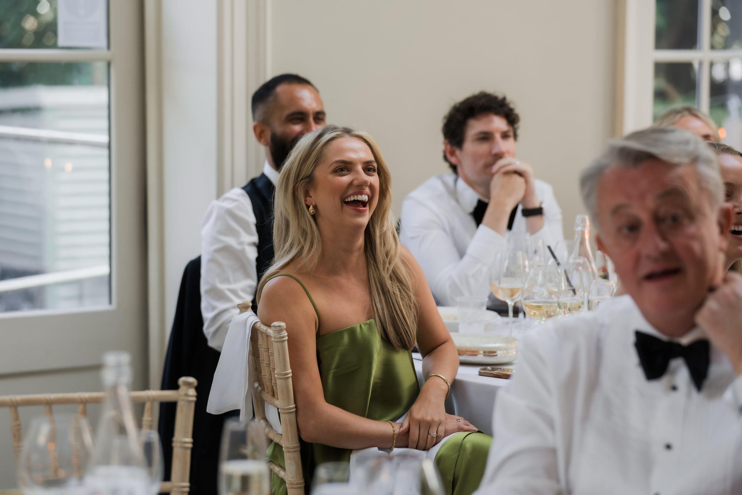 People sitting at a table during a formal event, laughing and enjoying themselves, with large windows in the background.