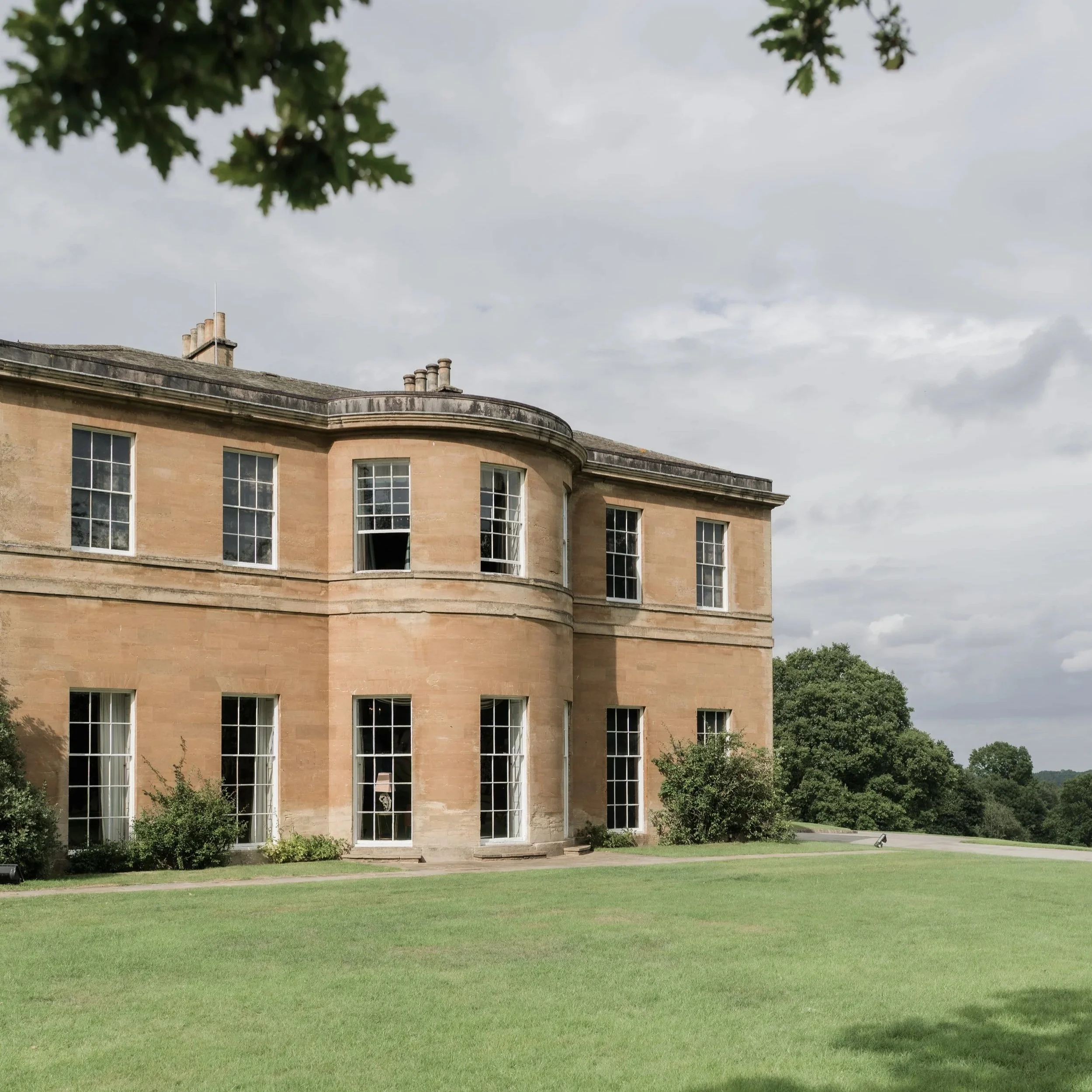 A large, historic building with beige stone walls and multiple tall windows, some open, in Yorkshire surrounded by a well-maintained lawn and trees under a cloudy sky.