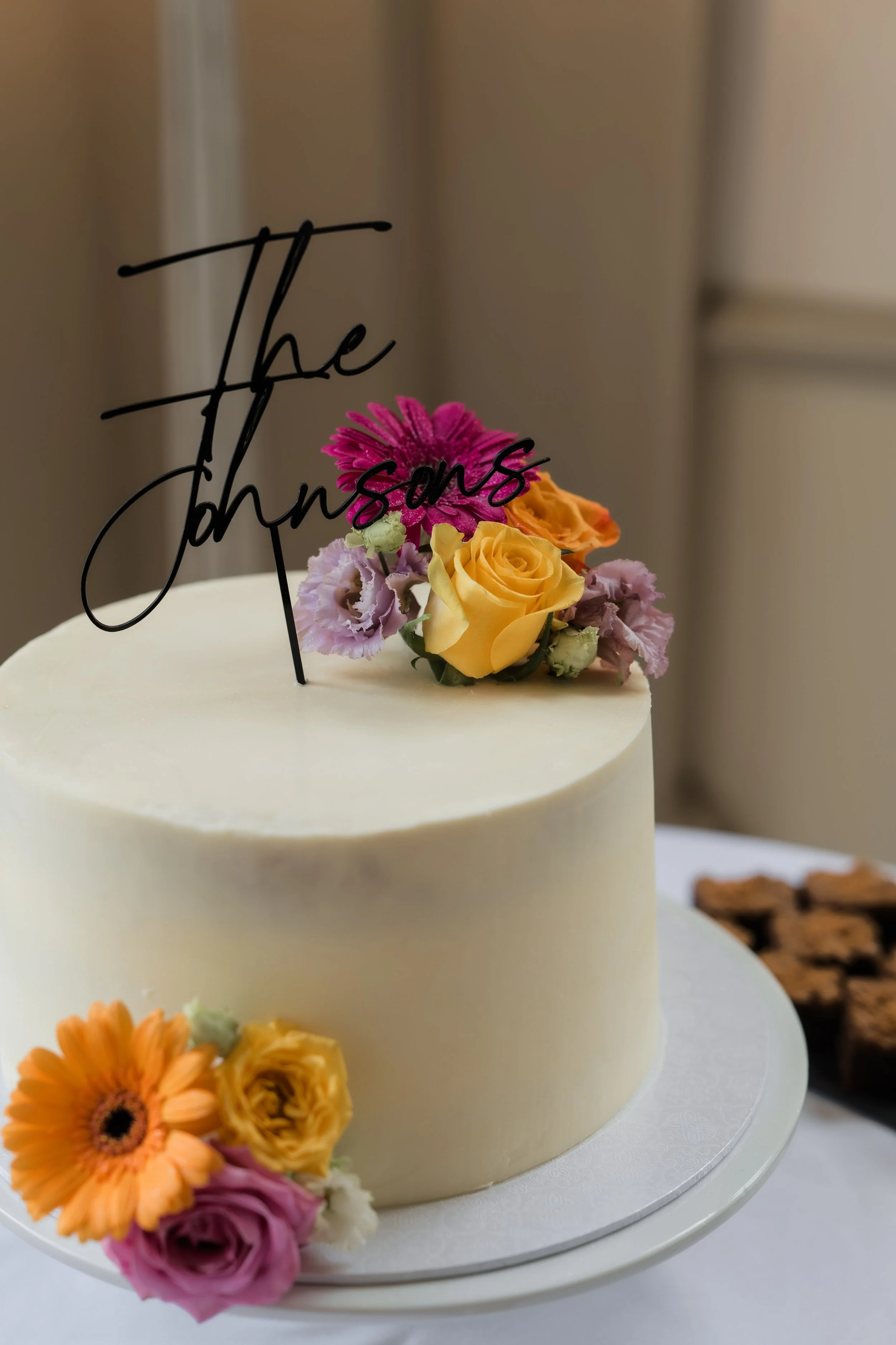A white frosted cake decorated with pink, yellow, and purple flowers, and a black cursive cake topper that reads 'The Johnston's'.