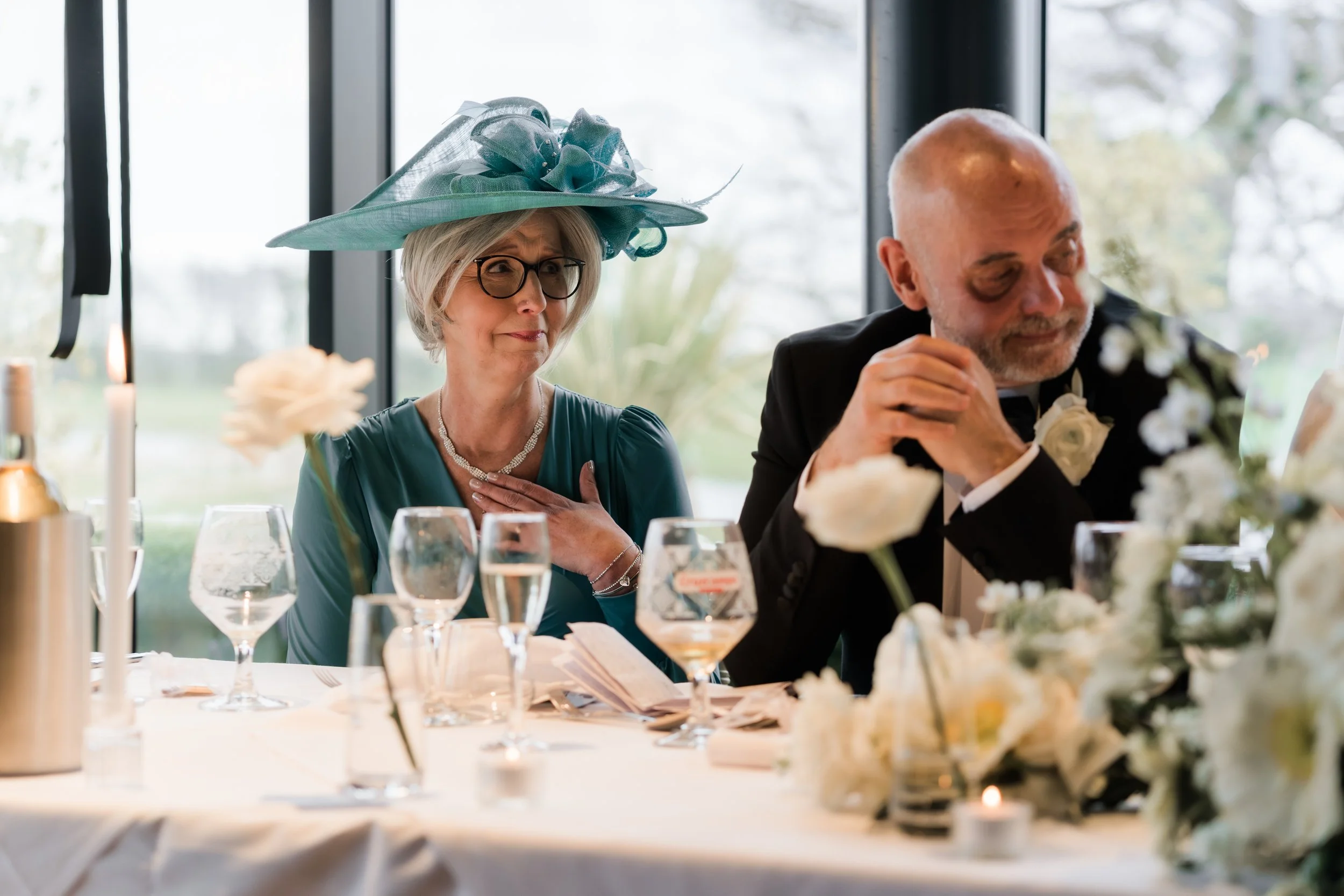 An elderly woman in a teal dress and large teal hat with netting sitting at a decorated table with a man in a tuxedo during a formal event, likely a wedding or celebration.