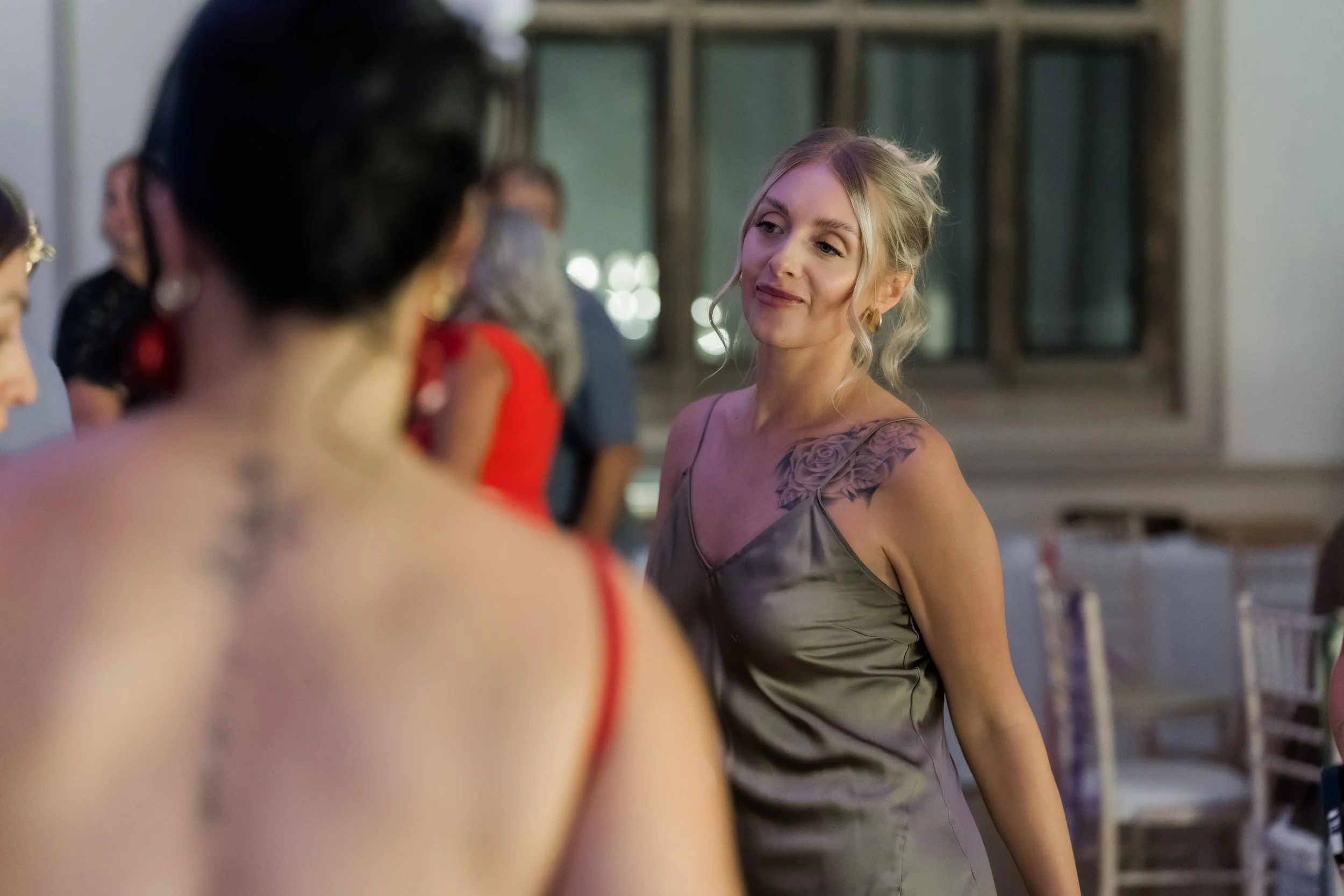A woman in a silver dress with a tattoo on her shoulder smiling while talking to another woman at a social event.