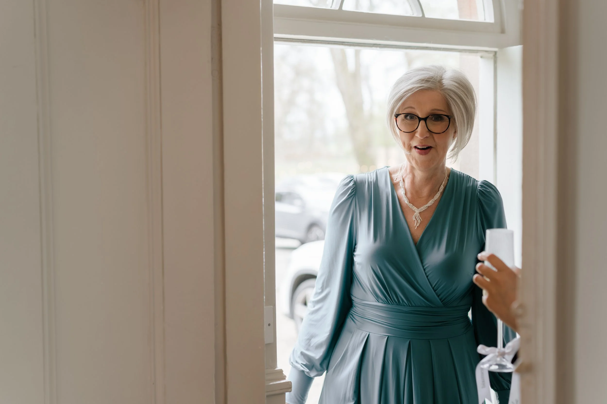 An elderly woman with gray hair and glasses standing at a doorway, wearing a teal dress and a pearl necklace, holding a small gift box with a white ribbon.
