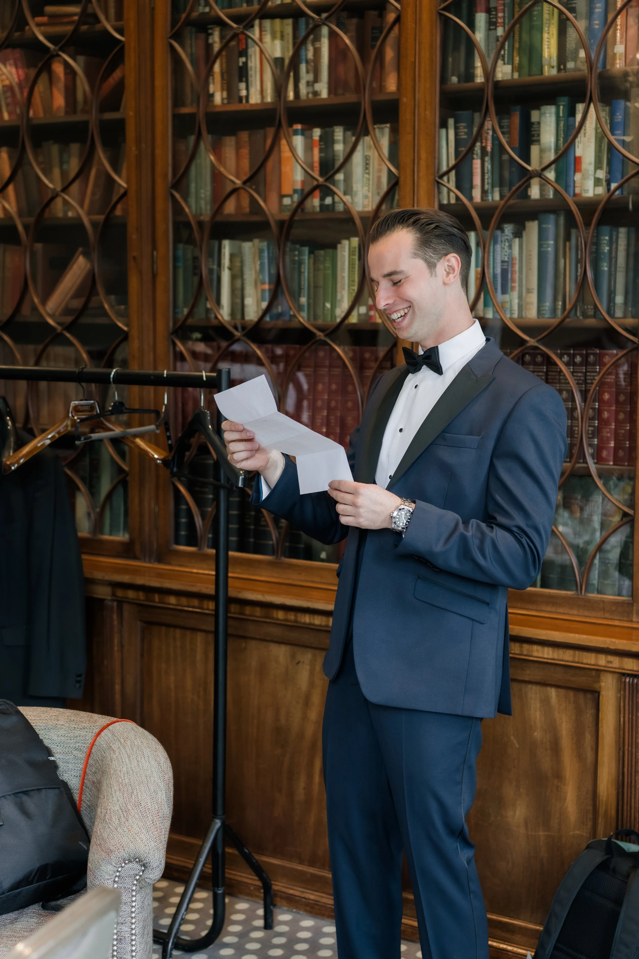A man in a tuxedo smiling while reading a piece of paper in a library with wooden bookshelves.