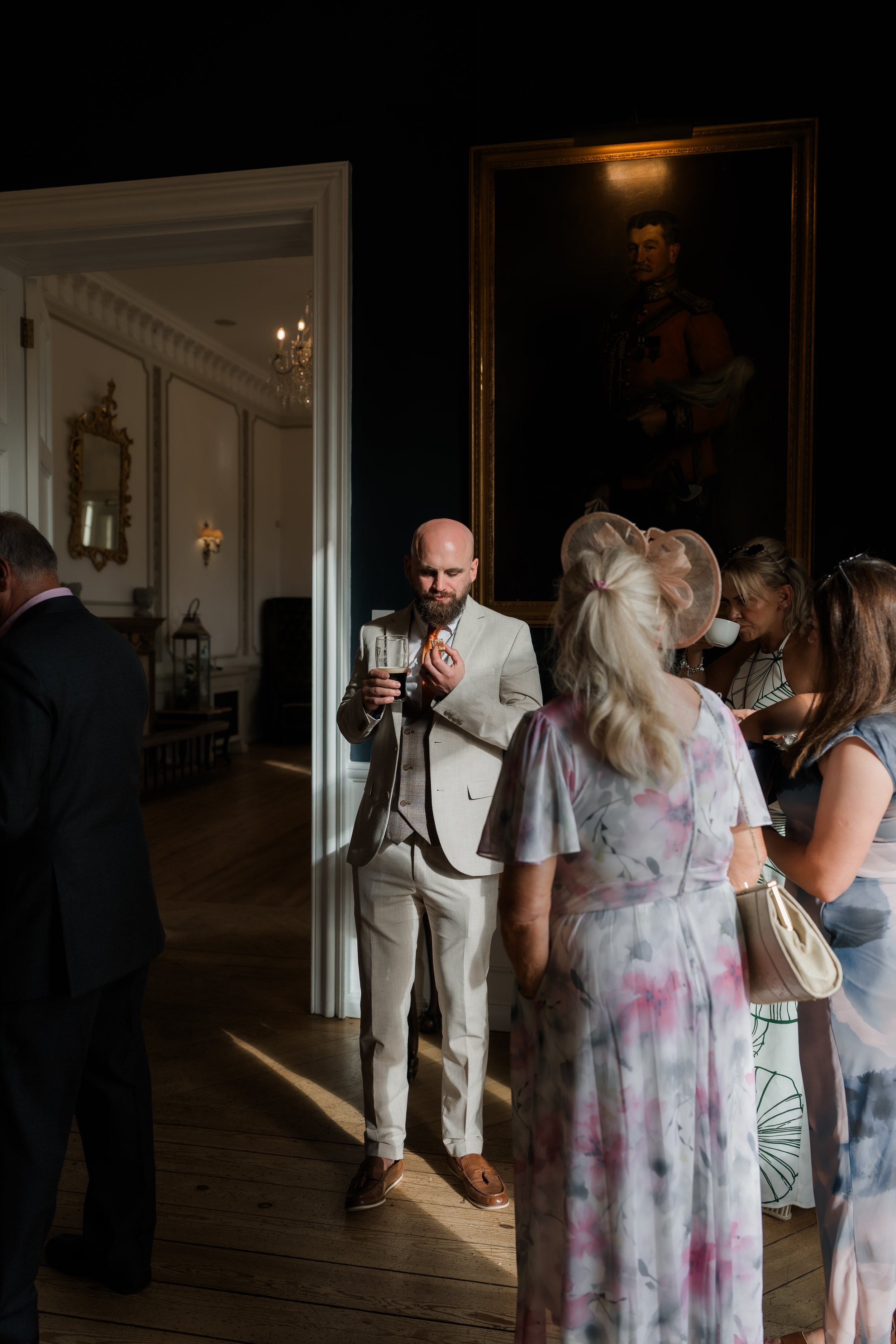 People socializing at an elegant indoor event, with a man in a light-colored suit holding a drink and eating, and women wearing hats and dresses chatting nearby, in a room with dark walls and a large portrait painting.