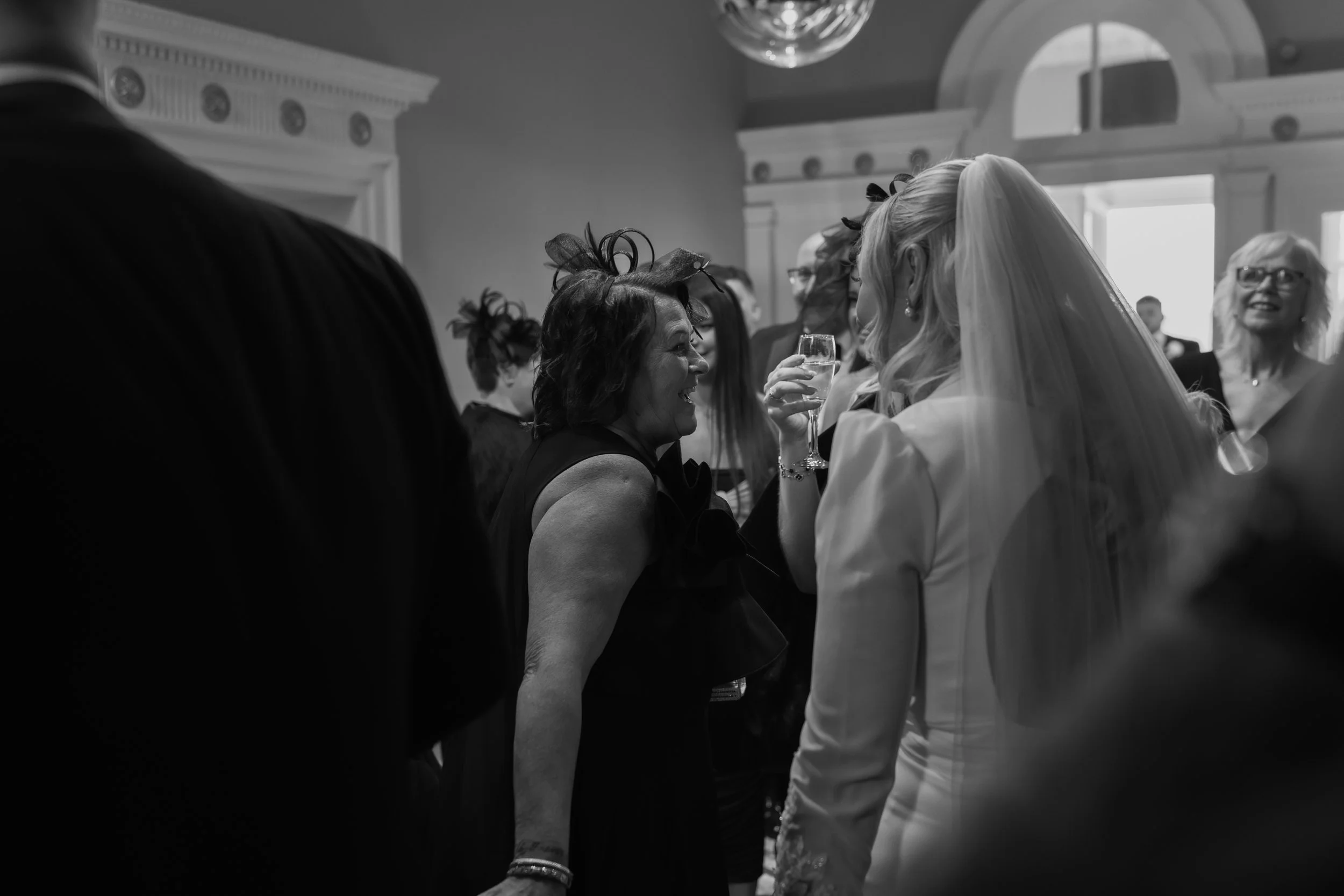 A black-and-white photo of a wedding reception with a bride in a white wedding dress and veil talking to a woman in a dark dress holding a glass of champagne, surrounded by other guests.
