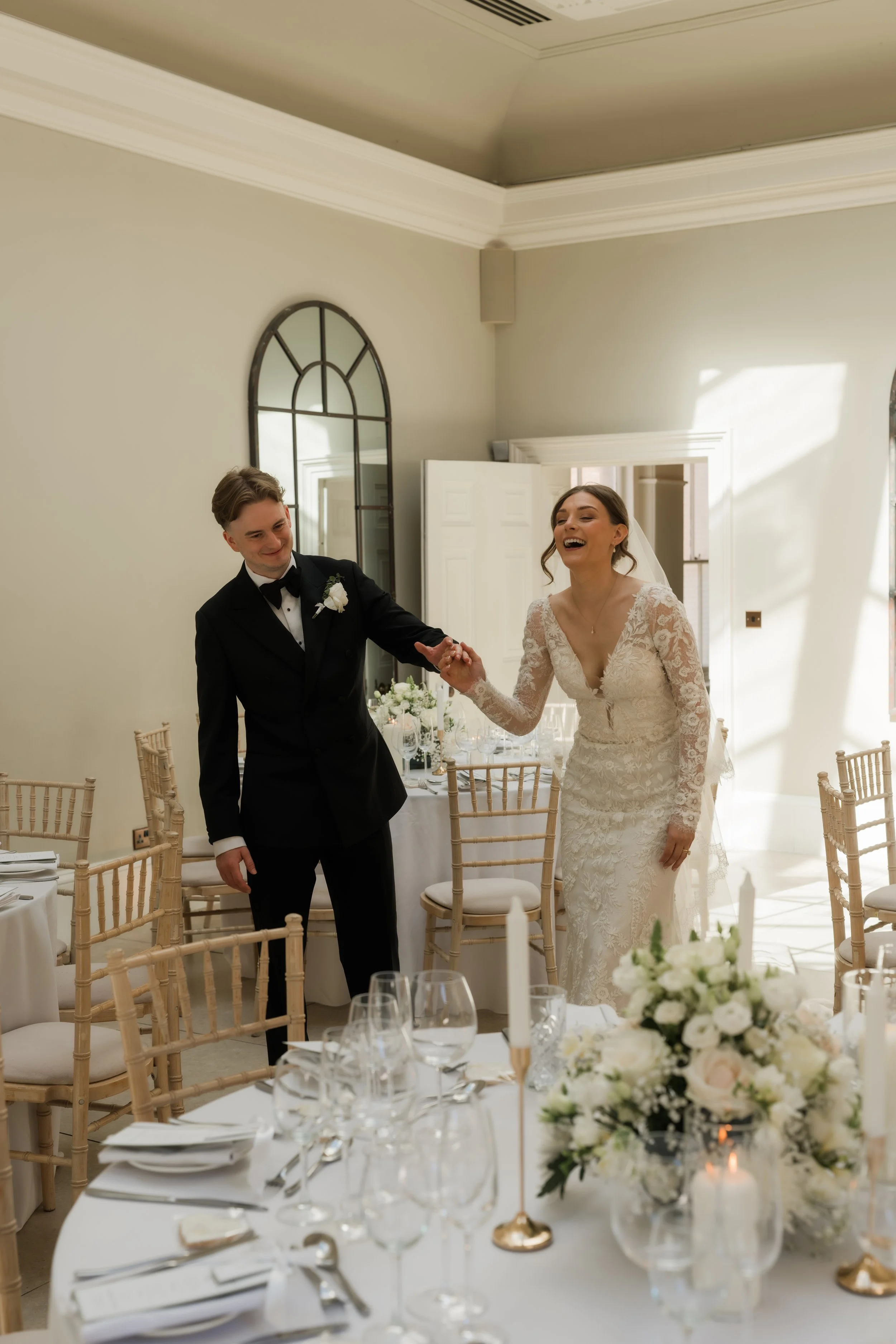 A bride and groom dancing at their wedding reception in a well-lit room with white walls and large windows. The bride is wearing a lace wedding dress and veil, and the groom is in a black tuxedo. Table settings with white tablecloths, floral centerpi
