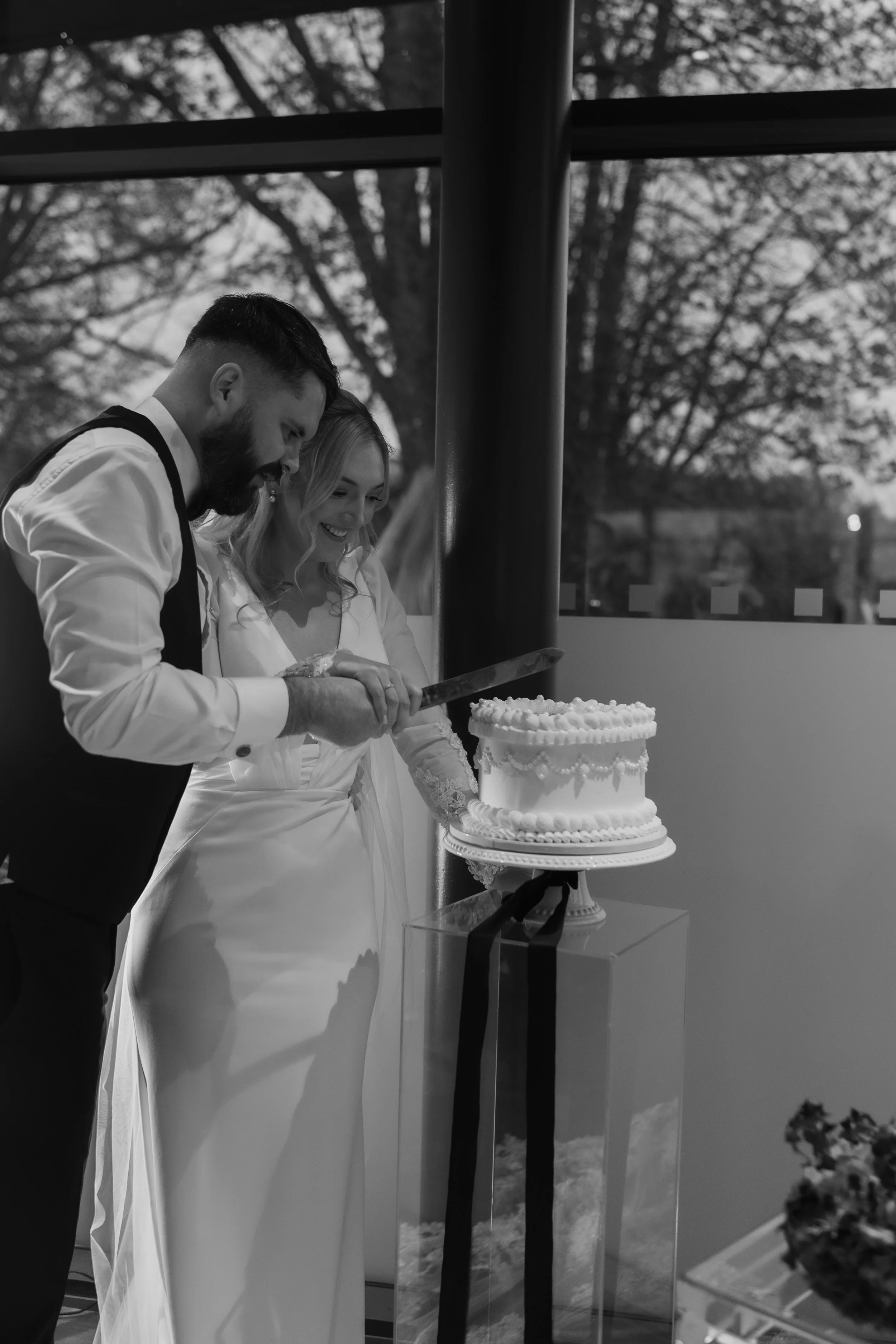 A black and white photo of a couple cutting a wedding cake, standing beside a large window with trees outside.