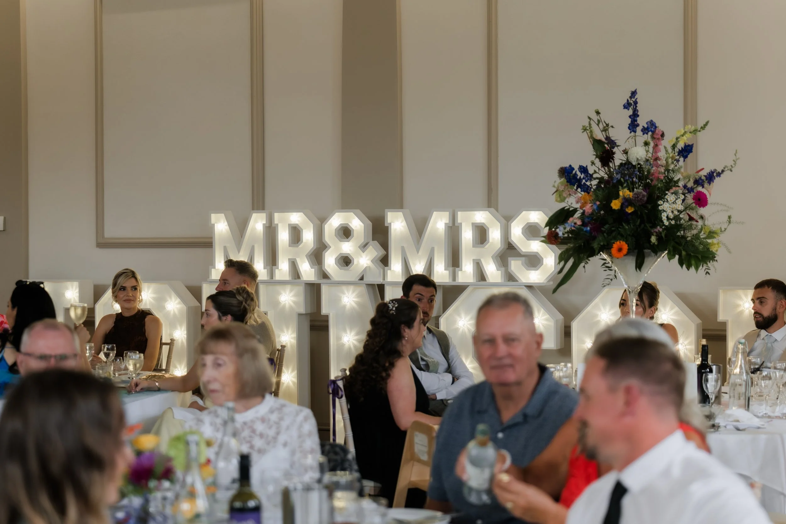 Wedding reception with guests seated at tables and large illuminated sign reading 'MR & MRS' in the background, decorated with flowers.