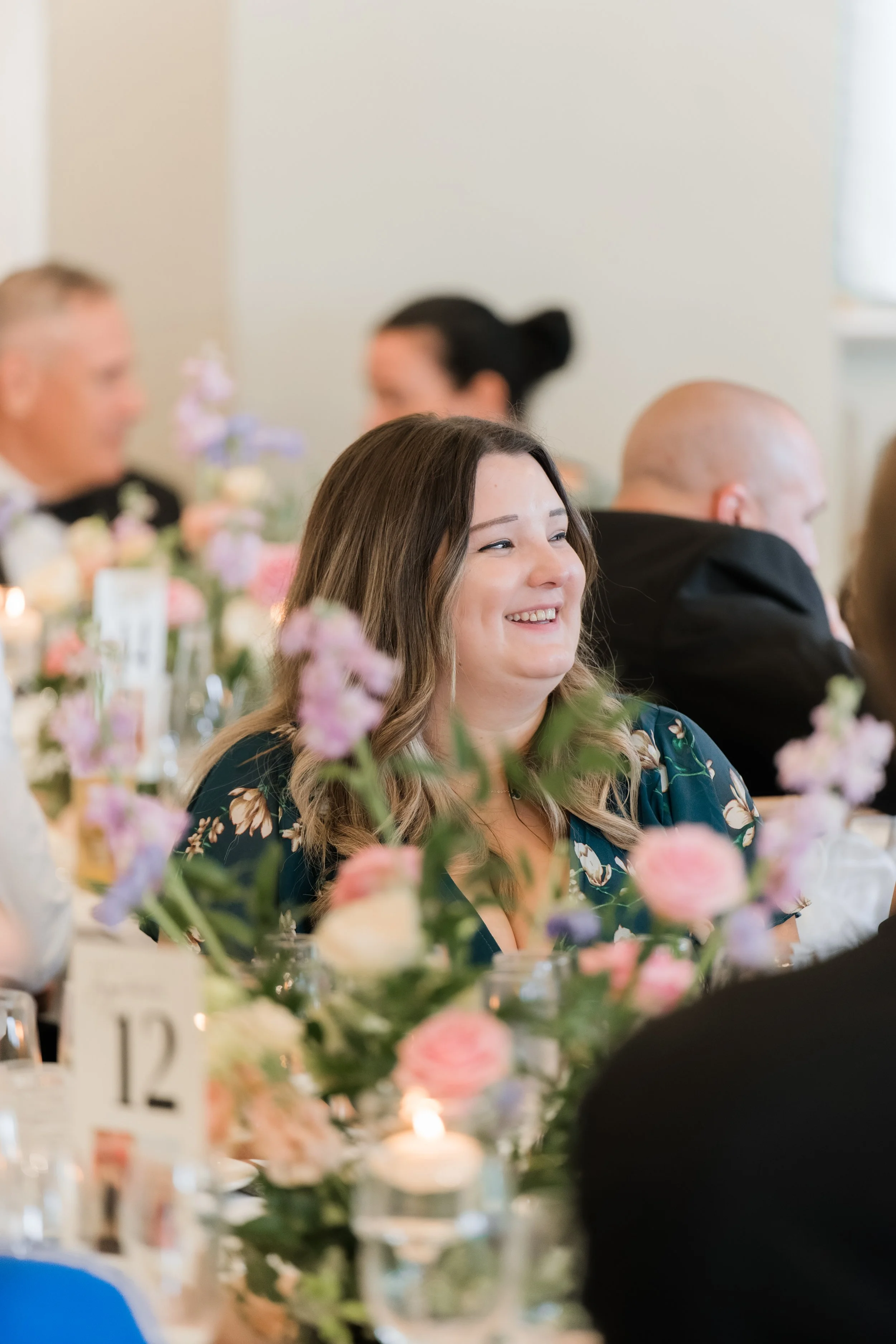 A woman smiling at a formal event, sitting among floral centerpieces with candles, at a table labeled with the number 12.