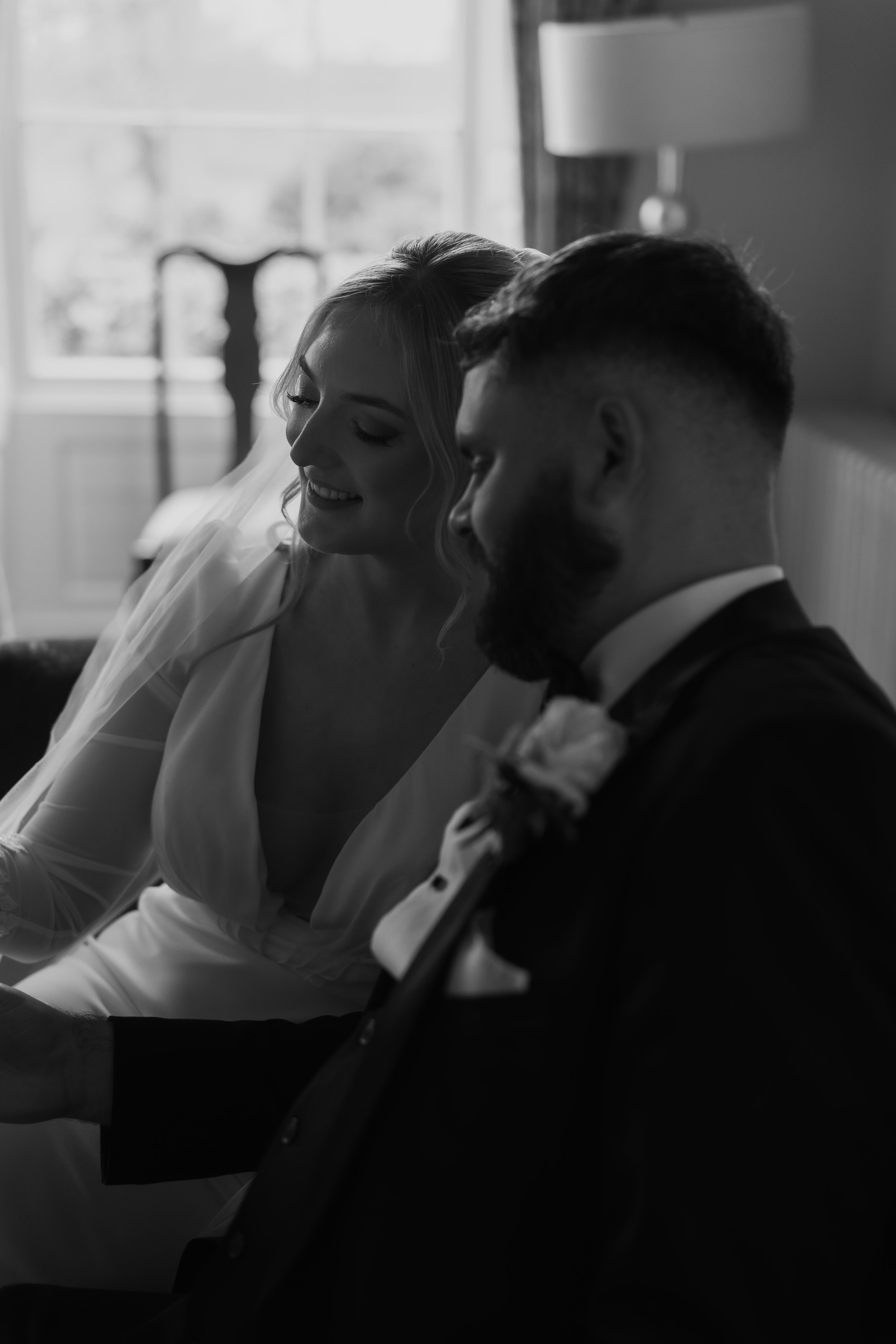 A black and white photo of a bride and groom sitting closely together indoors, smiling and sharing a tender moment.