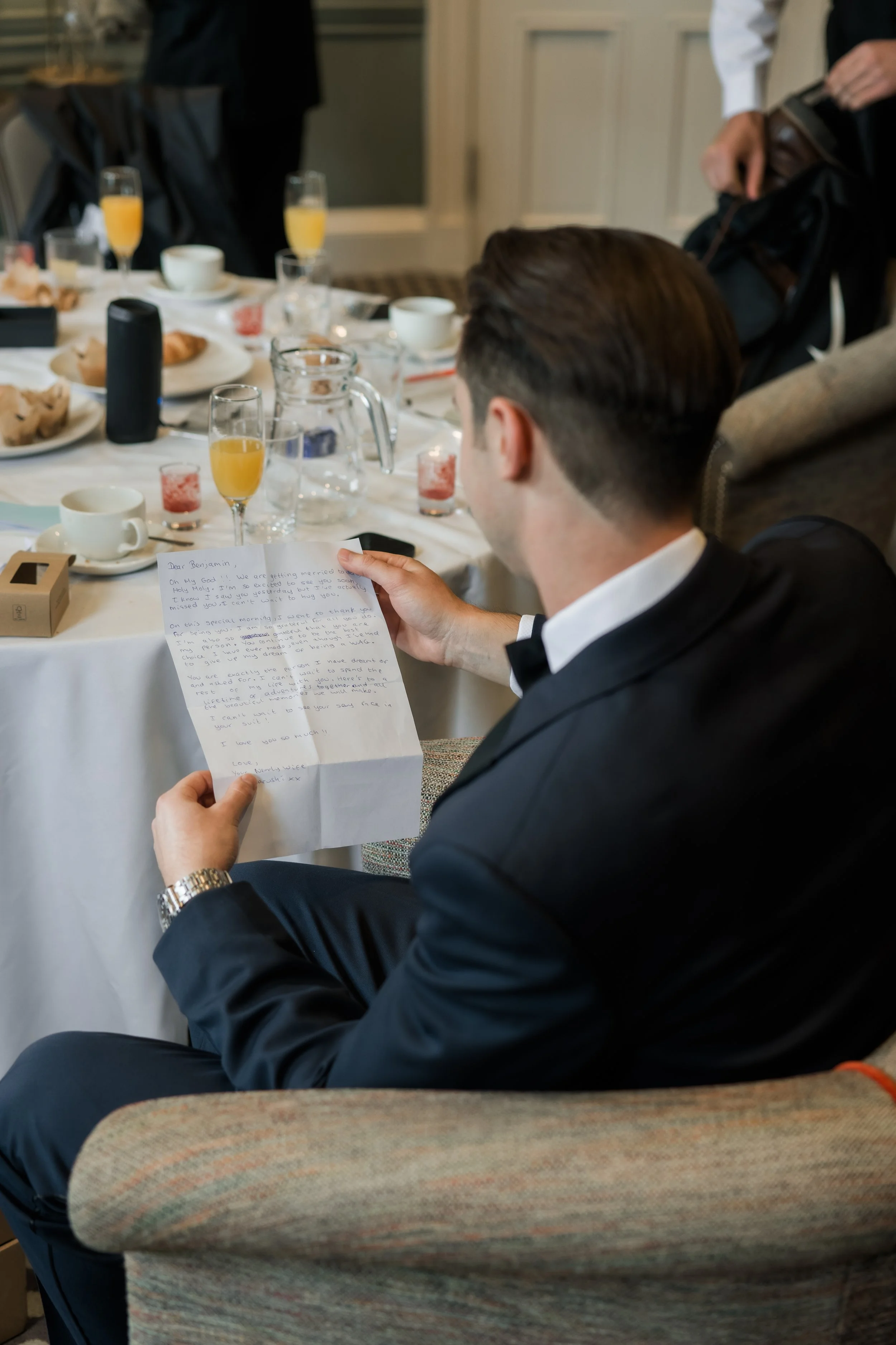 Man in tuxedo reading a handwritten letter at a formal gathering with a table of food and drinks.