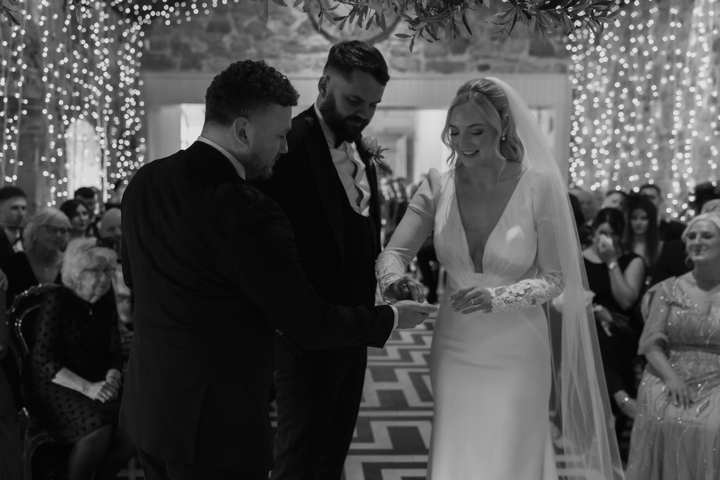 A black and white photo of a wedding ceremony with a bride and groom exchanging rings while a man officiates, in front of seated guests, with decorative lights in the background.