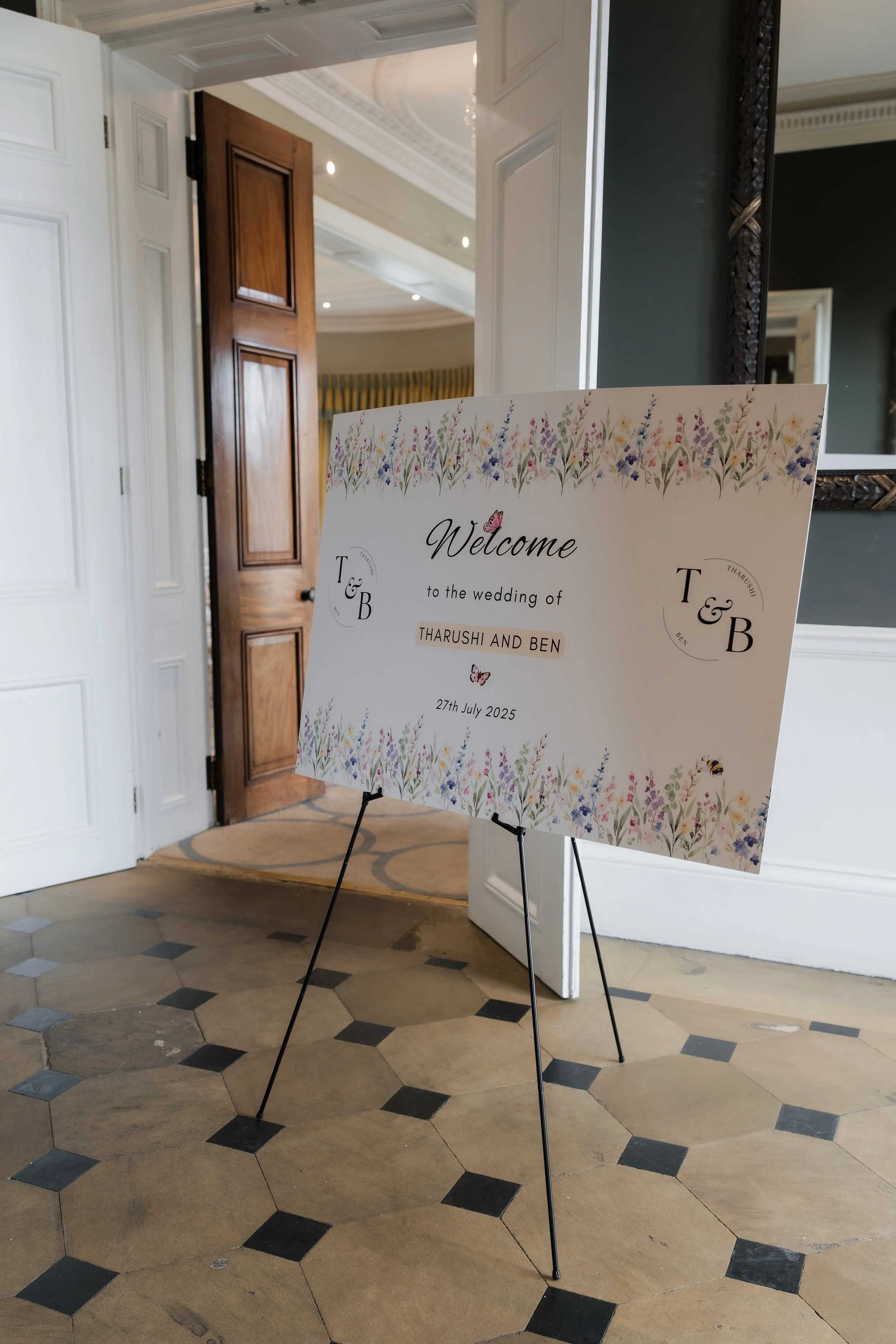 Wedding welcome sign with floral border, displaying the names Tharushi and Ben along with the wedding date, July 27, 2025, situated in an elegant interior with tiled floor and decorative ceiling.