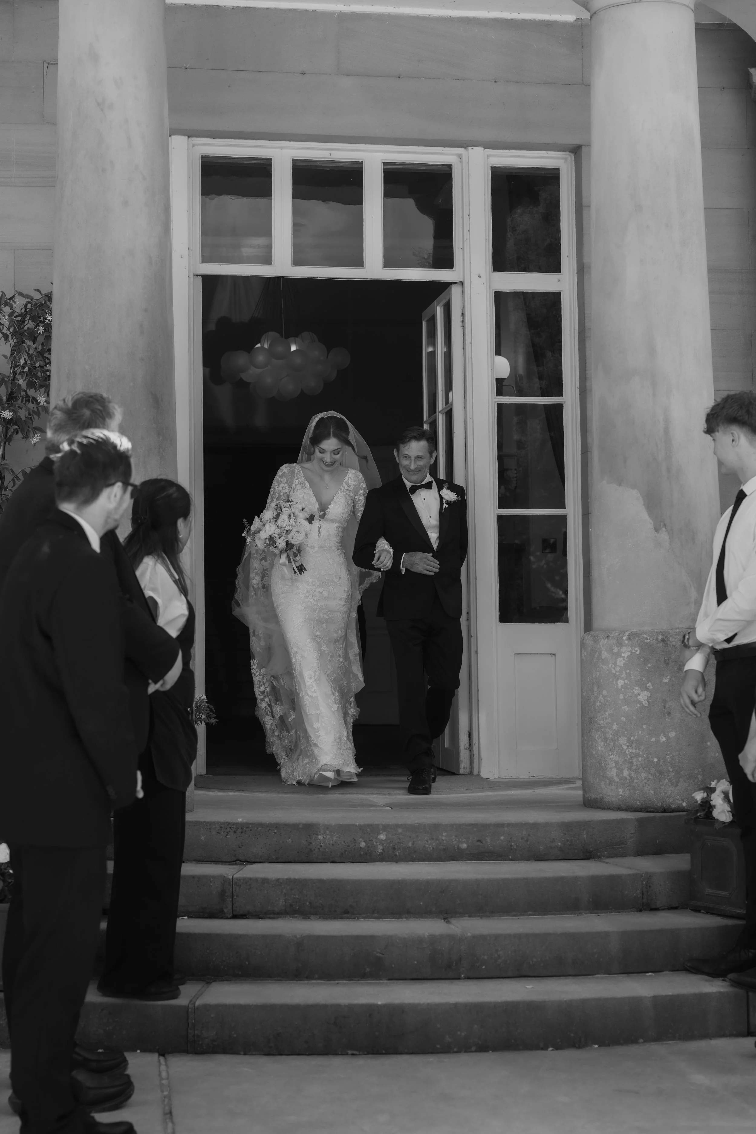 A bride in a wedding dress and a groom in a tuxedo walking down the steps of a building, surrounded by wedding guests dressed in formal attire.