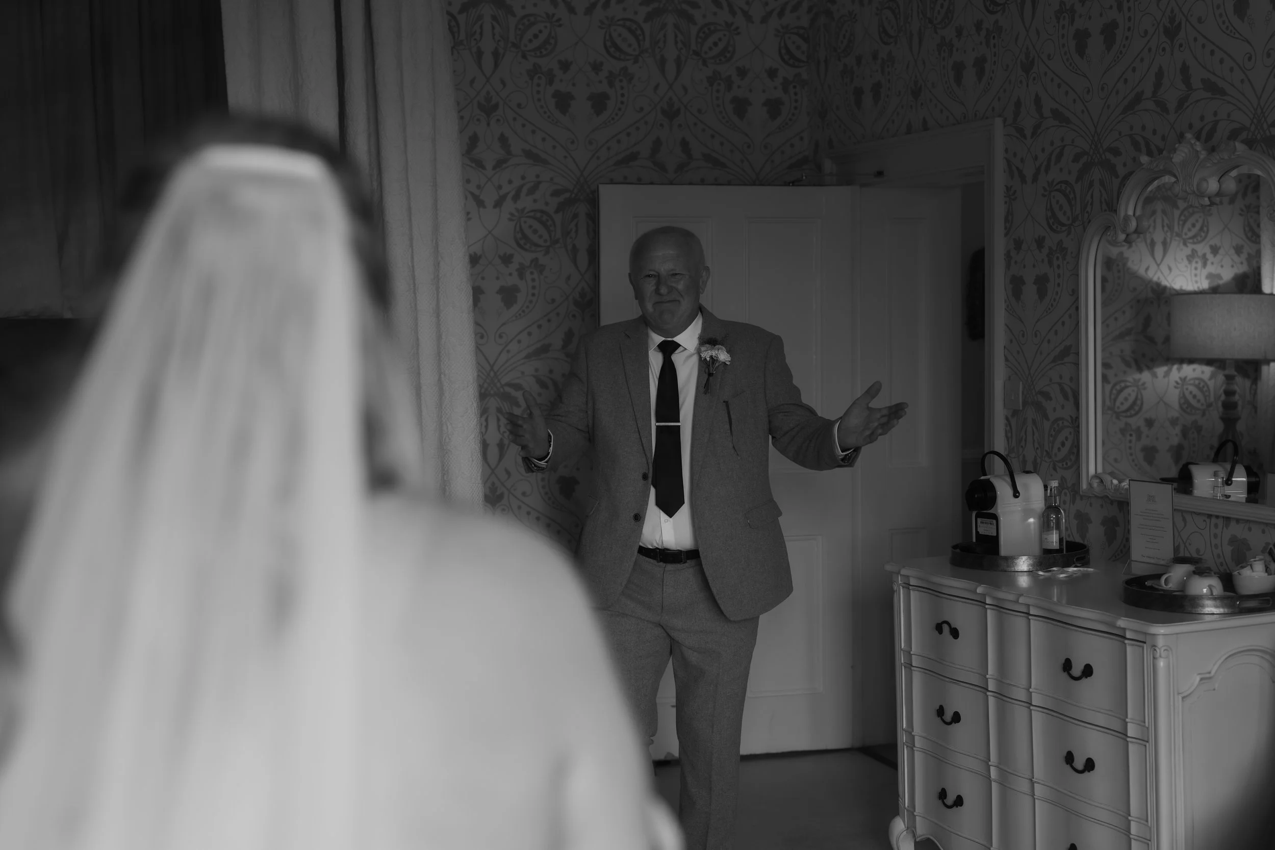 A man dressed in a suit with a boutonniere is smiling and gesturing with open hands in a room, with a woman in a veil and wedding dress in the foreground.
