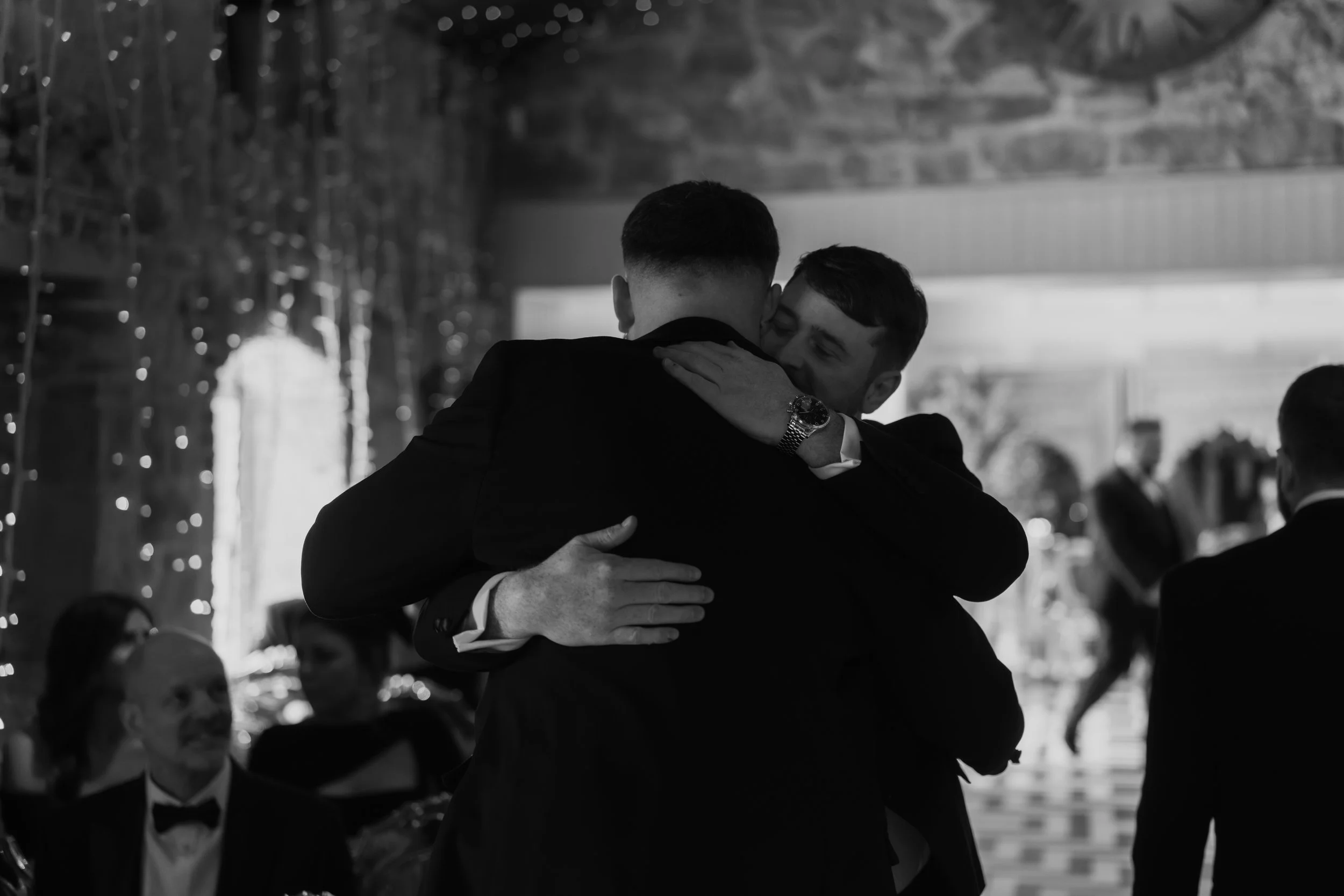 Two men in formal attire hugging at a gathering or event, with other people seated and talking in the background.