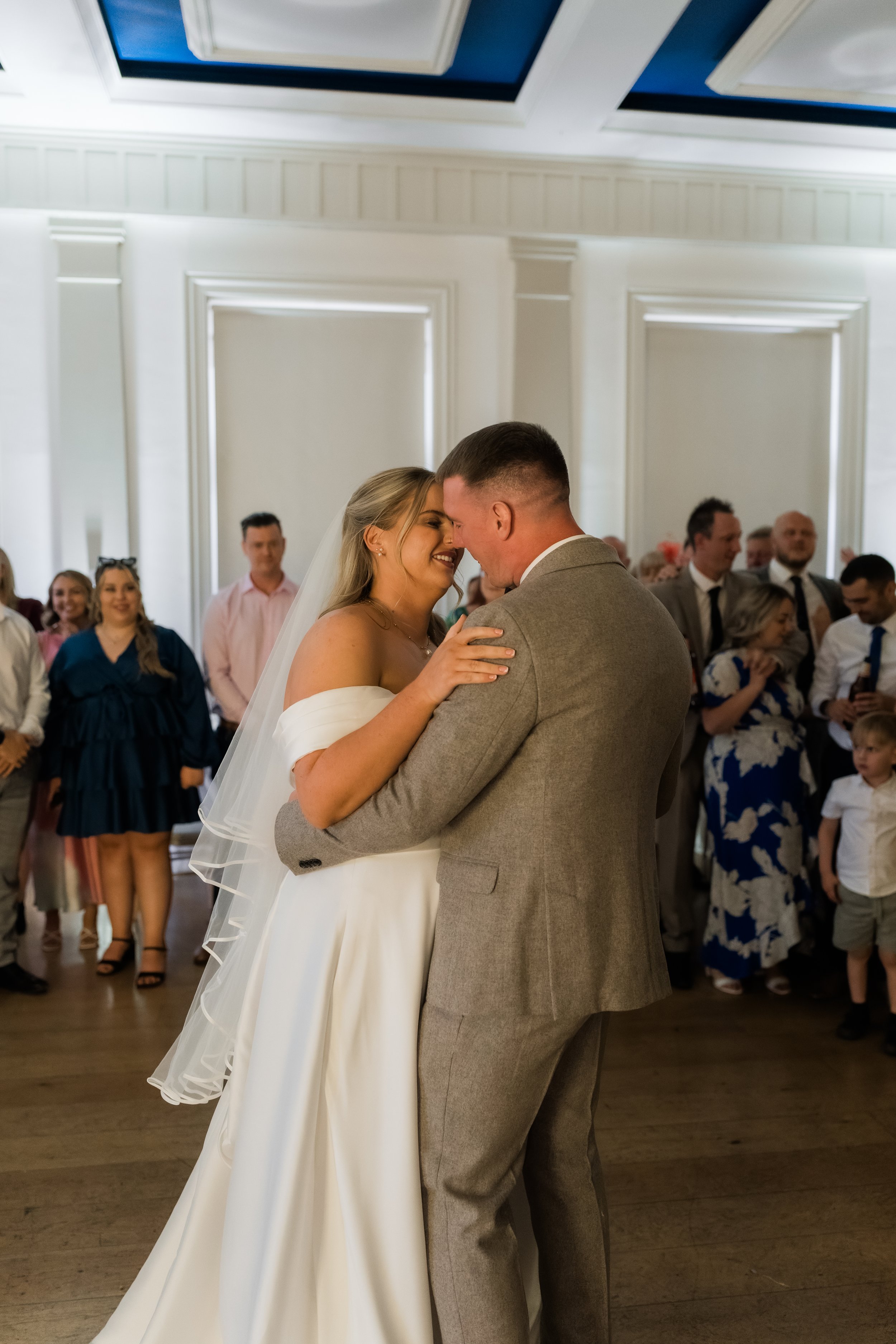 A bride and groom share a dance at their wedding reception, with guests watching and smiling in the background.