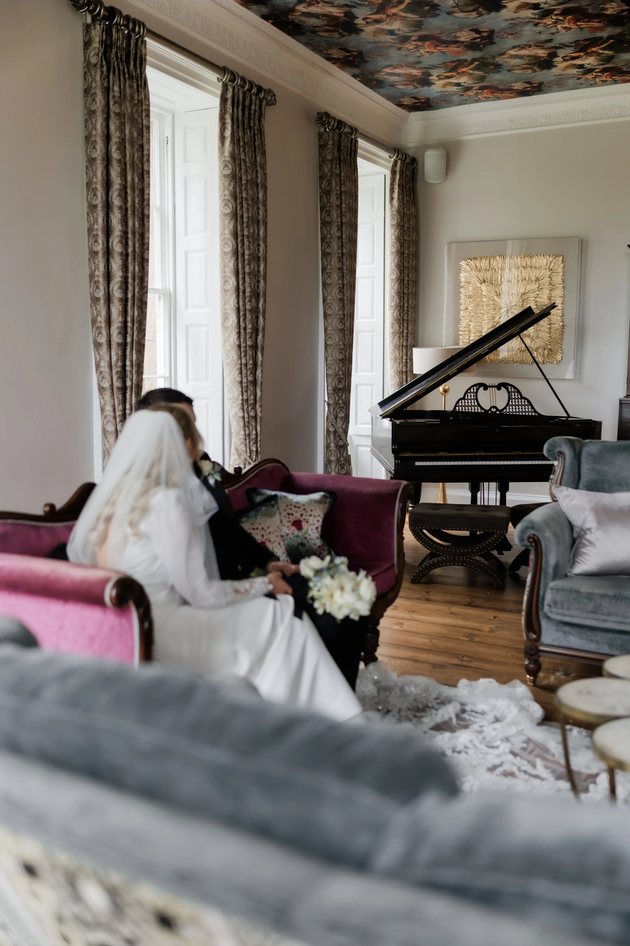 A bride dressed in white sitting on a pink vintage sofa, holding a bouquet of white flowers, with a groom in dark attire sitting beside her, in a well-lit formal living room with tall windows, beige walls, patterned curtains, a black grand piano, and