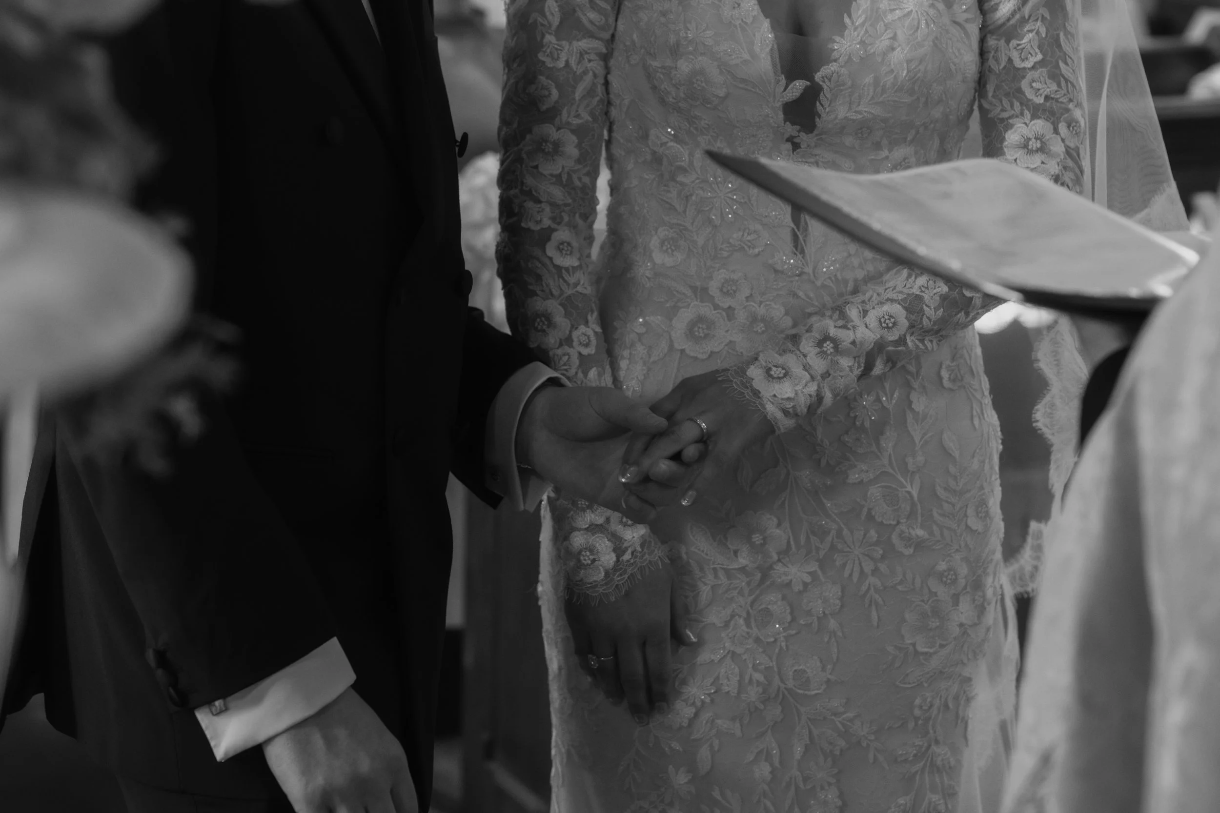 A black and white photo of a wedding ceremony, showing the bride and groom holding hands. The bride is wearing a lace wedding dress and holding a book, while the groom is dressed in a dark suit with a white shirt.