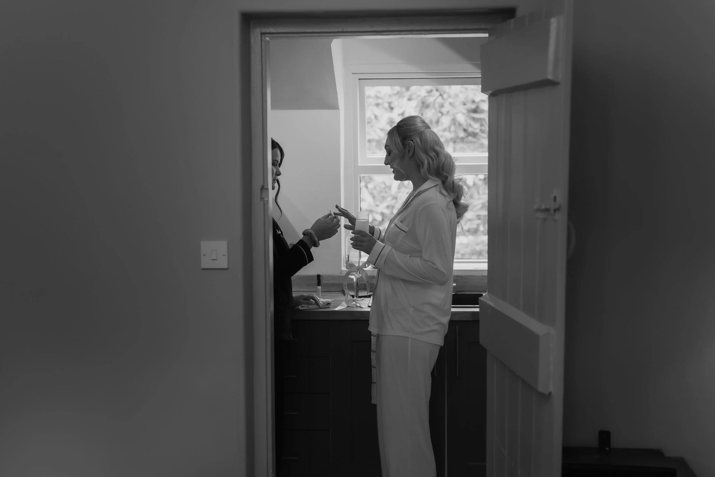 Two women standing in a kitchen, one wearing pajamas and the other in dark clothing, having a conversation, with the woman in pajamas holding a glass of milk.