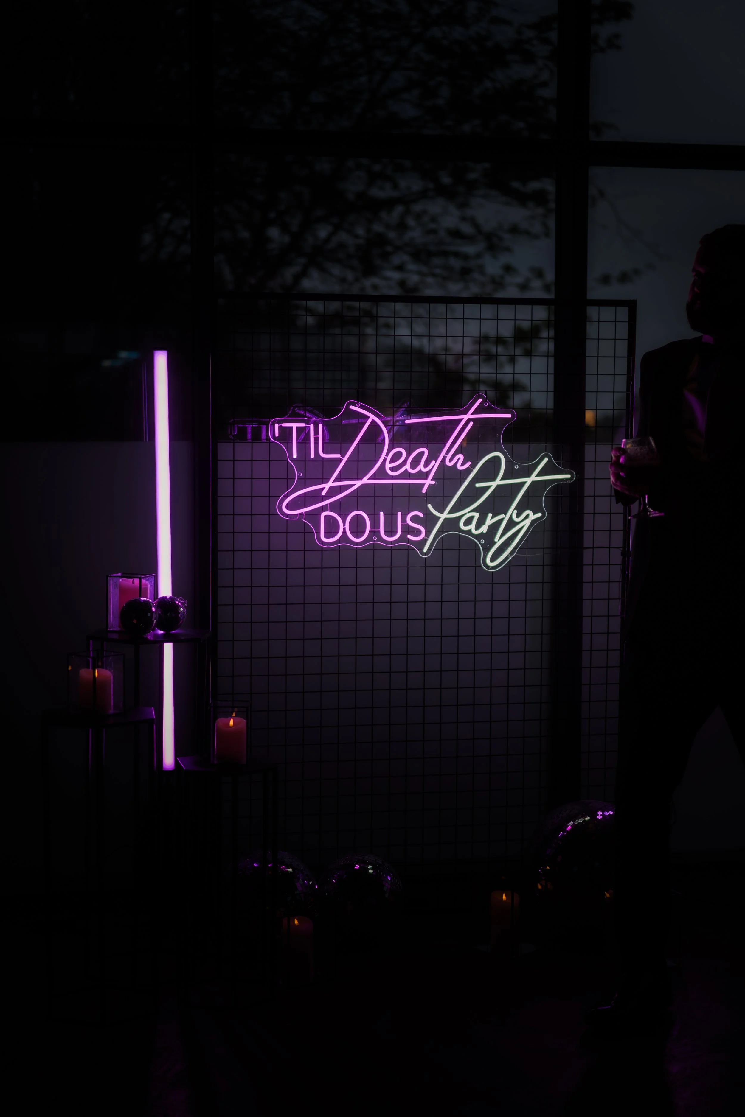 Neon sign reading 'Still Death Does Part' in pink and green lights, with candles and decorative objects on black tables, and a man holding a drink on the right, with a window showing a dark sky and trees outside.