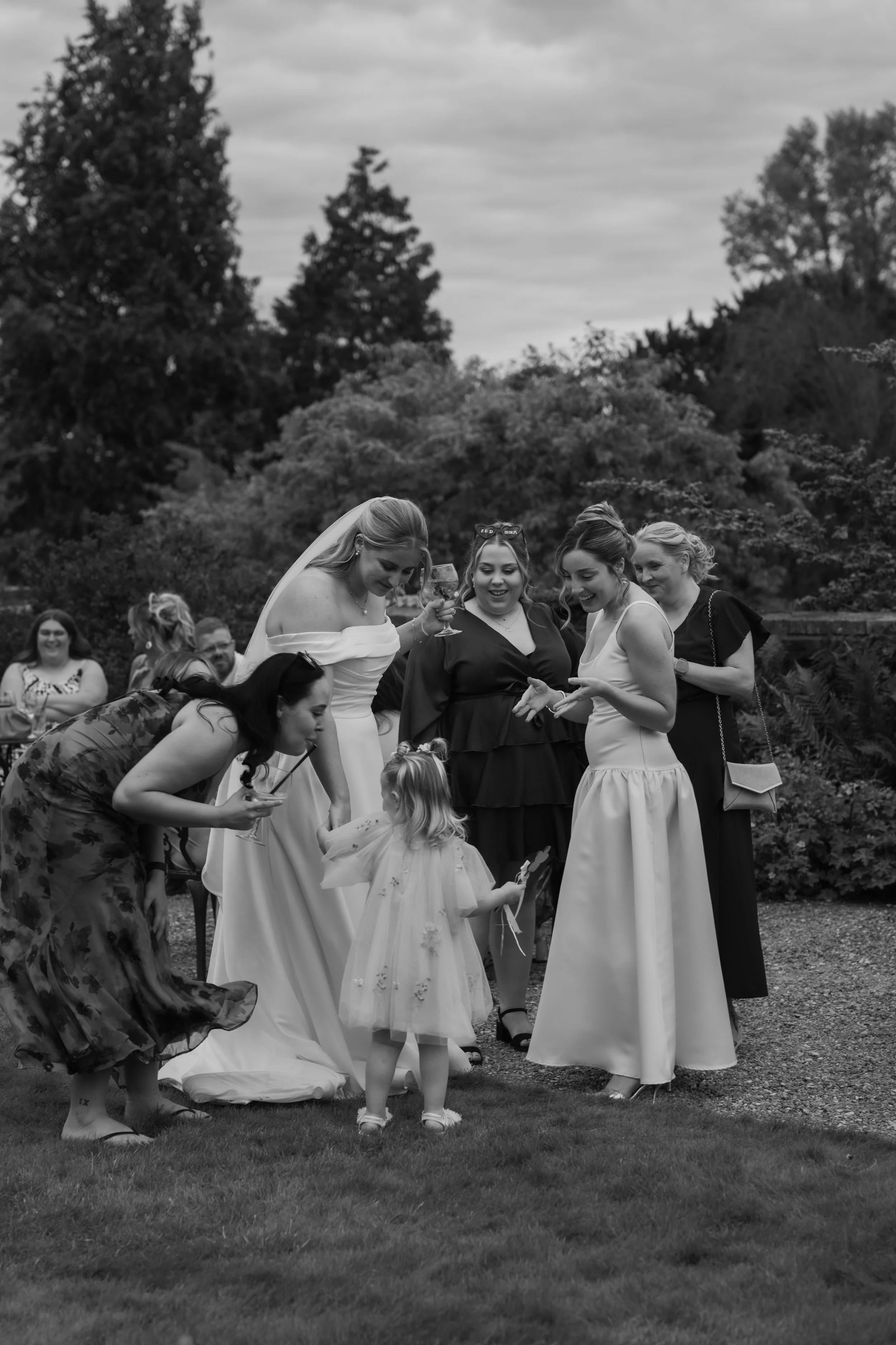 A group of women, including a bride in a wedding dress, and a young girl, gathered outdoors on grass, smiling and looking at something the girl is holding, with trees and others seated in the background.