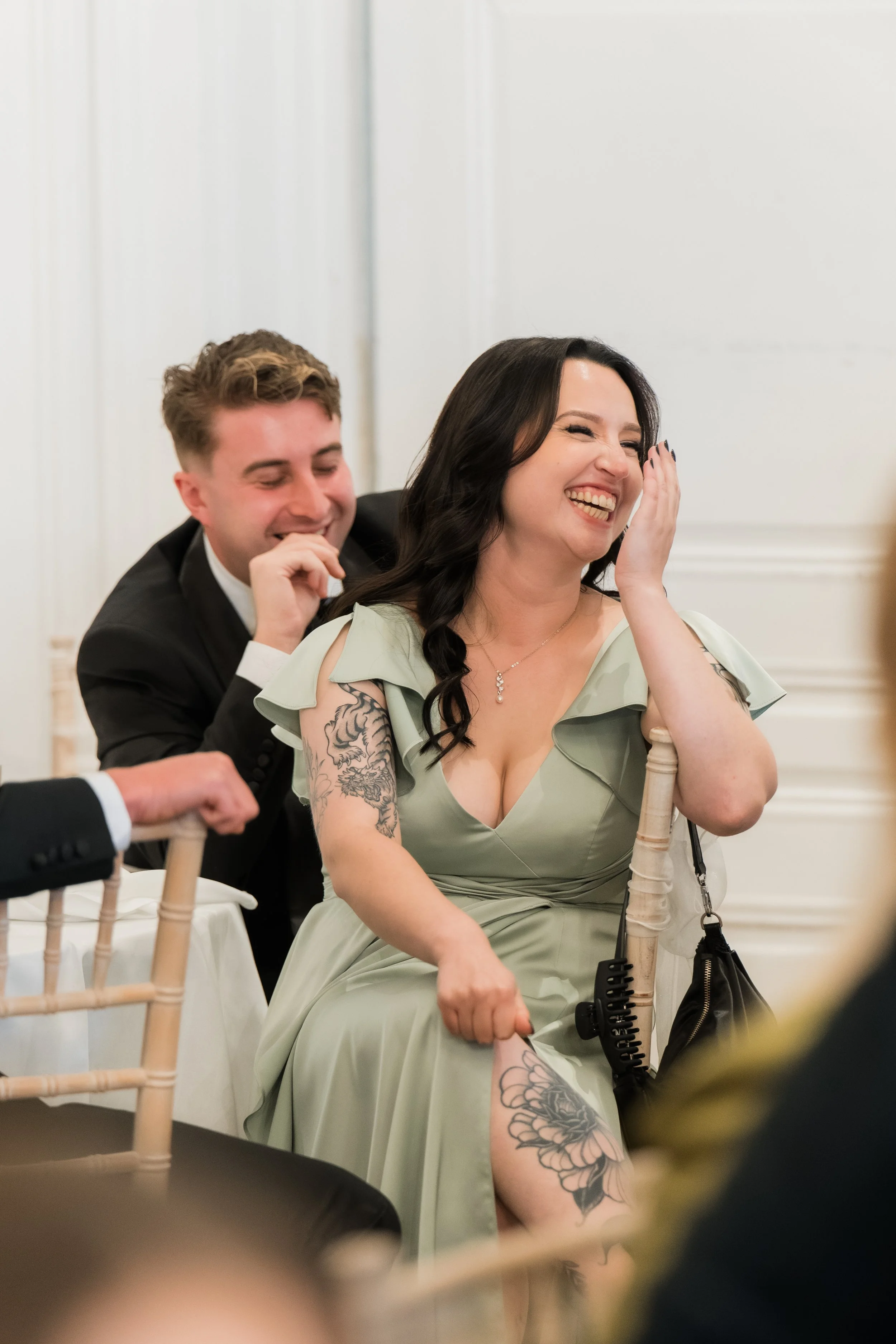 A woman in a light green dress laughing and covering her face while a man in a black suit behind her also laughs, sitting on a white chair at a formal event.