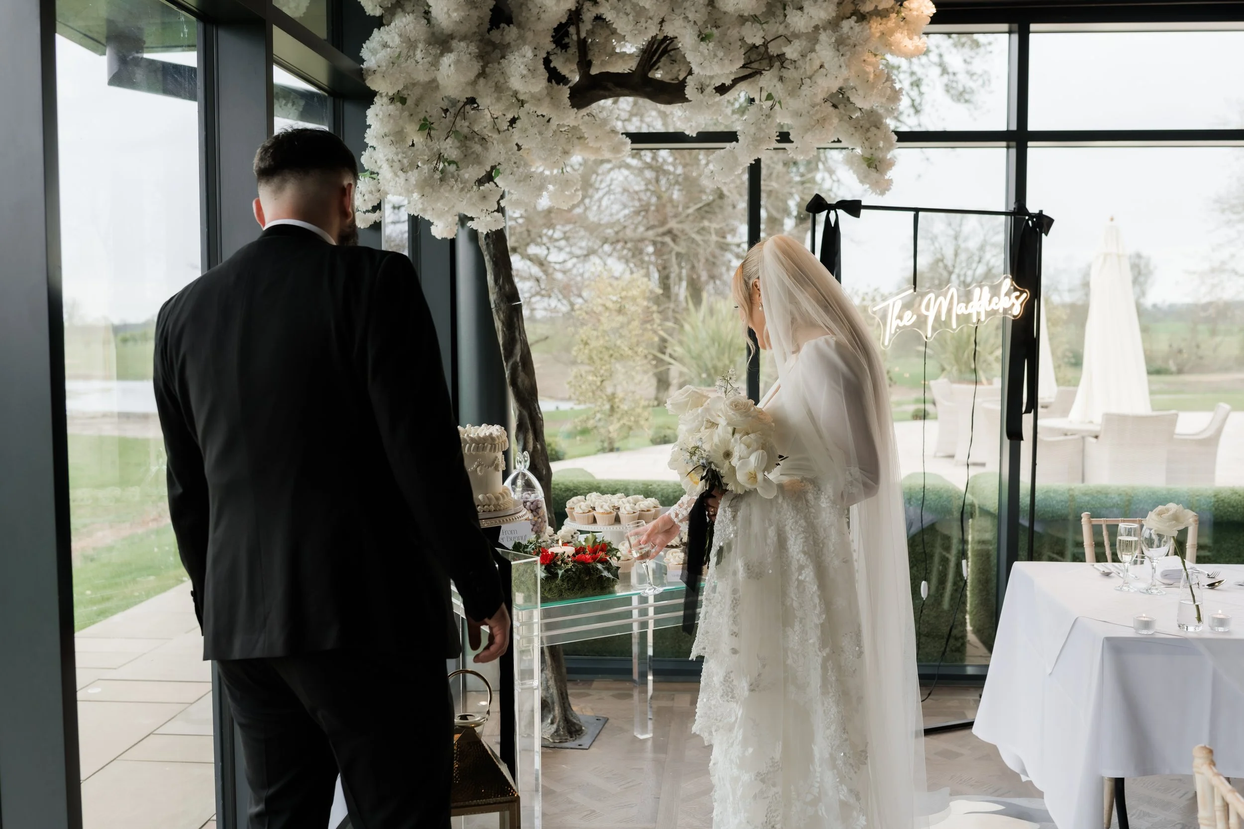 Bride and groom at wedding reception, bride in white lace dress with veil, holding bouquet, standing near decorated table with flowers and desserts, large window with scenic outdoor view in background.