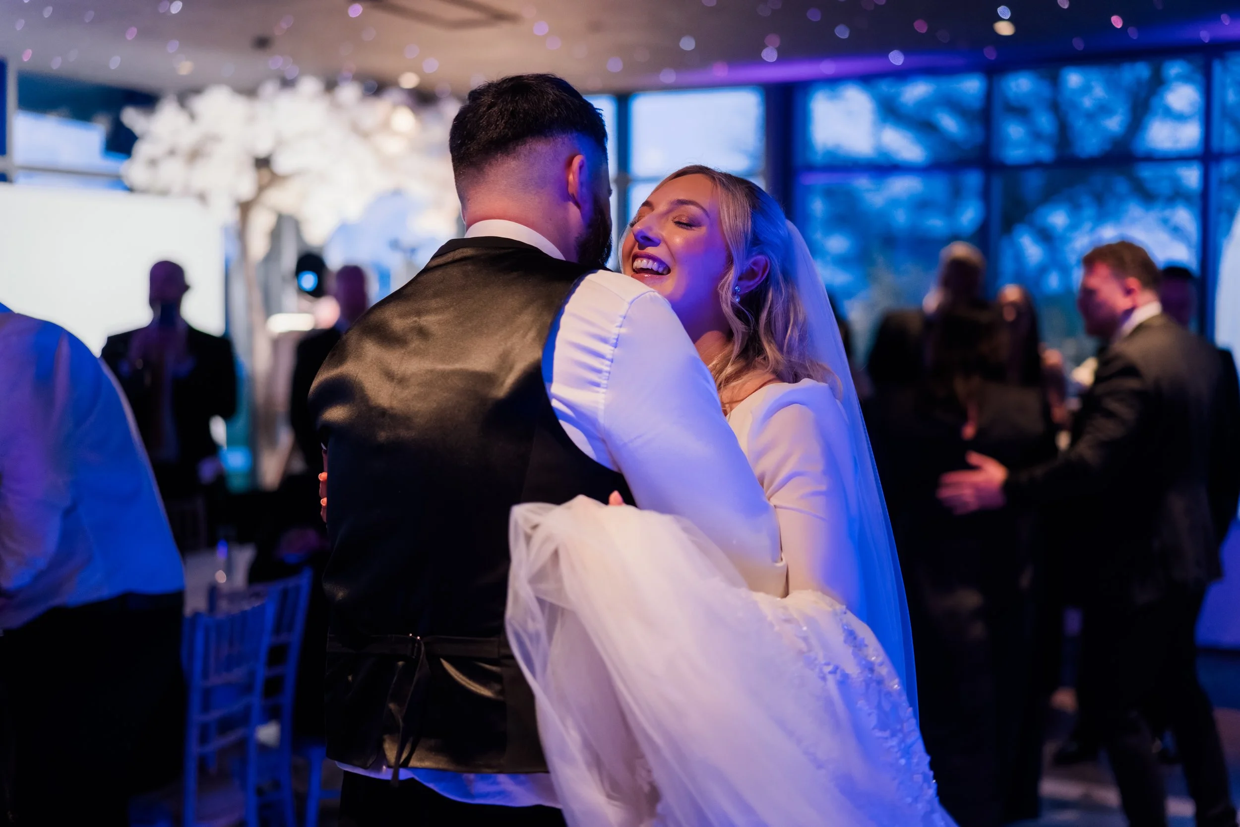 A bride and groom dancing at their wedding reception, smiling and embracing each other, with guests dancing in the background.