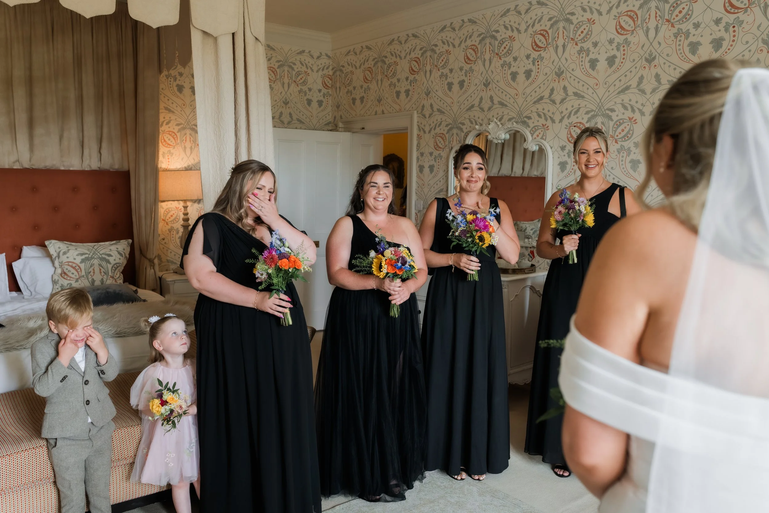 Bridal party and children during a wedding in a decorated room, with women holding bouquets and children in formal attire.