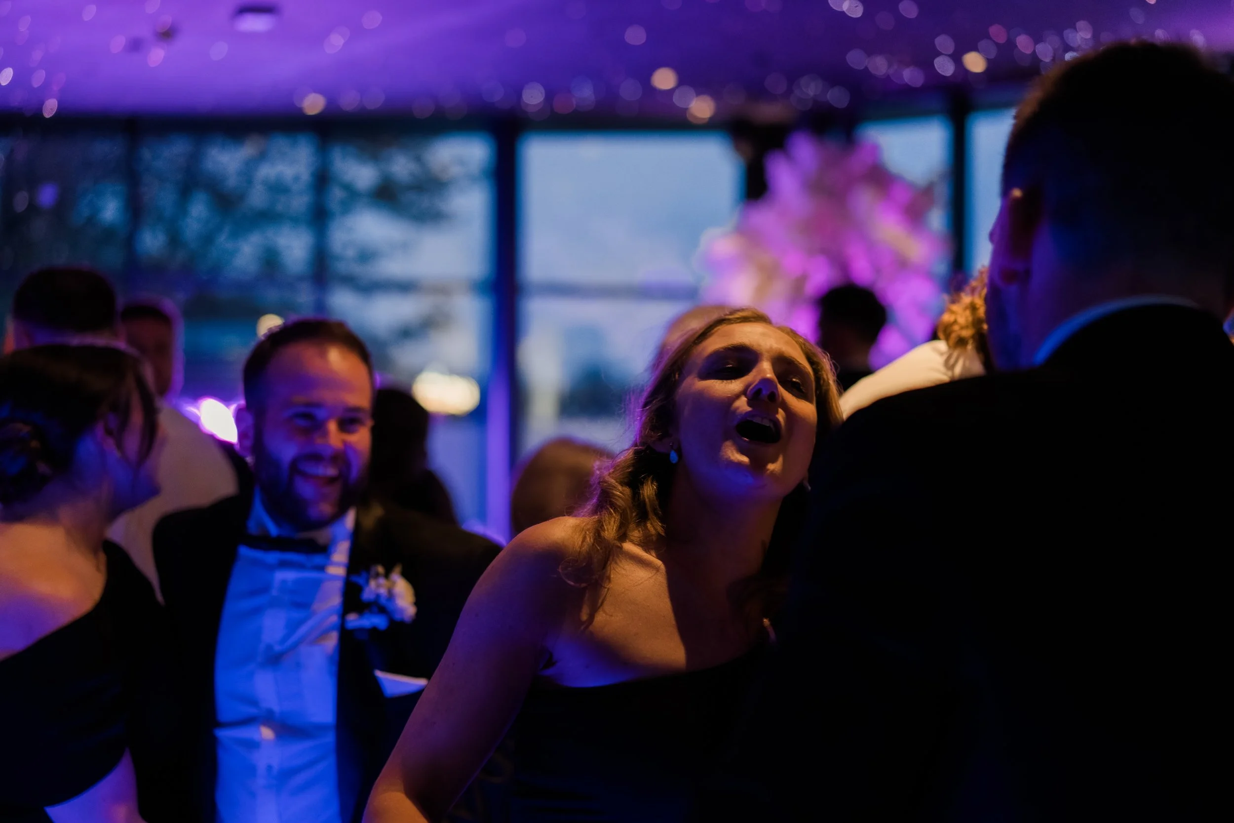 People dancing and socializing at a wedding reception with purple lighting and large window in the background.