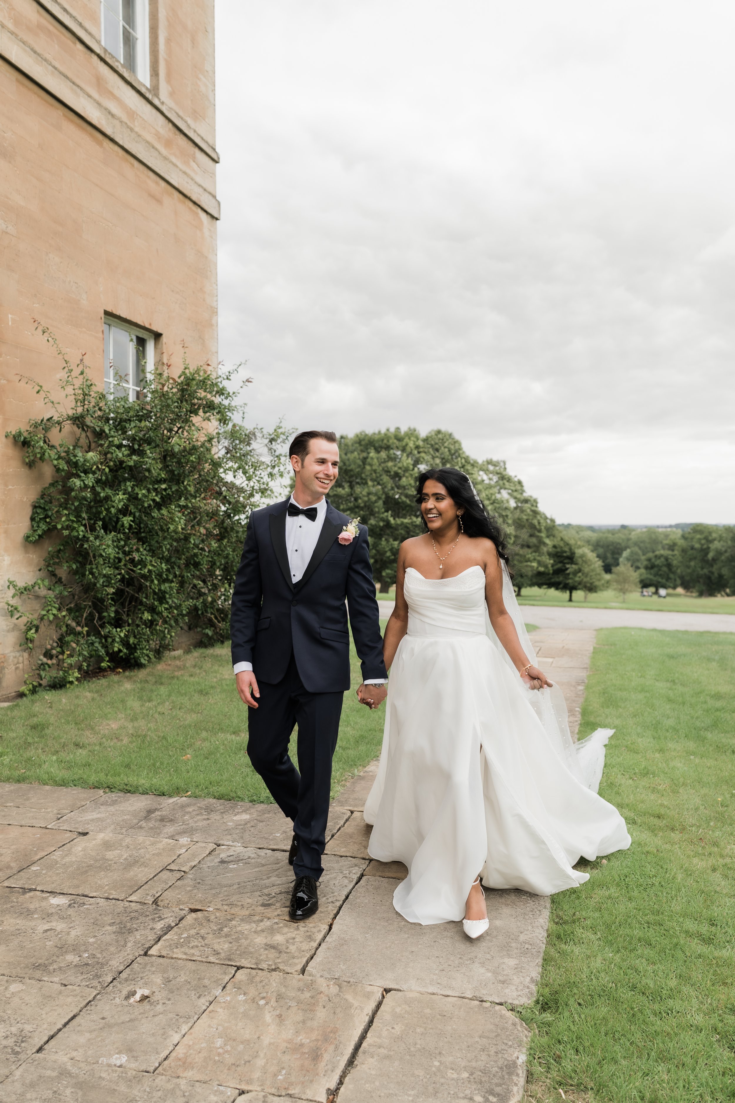 A happy bride and groom holding hands and walking outside on a cloudy day, dressed in wedding attire, near a stone building and green landscape.